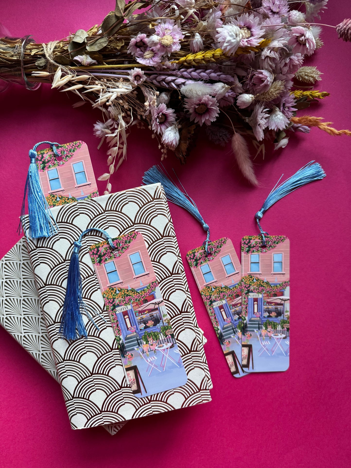 A coffee shop illustrated bookmark with a blue exterior, an outside table and umbrella and building windows surrounding. This illustration is filled with lots of plants and flowers and features cups of coffee and baked goods. Illustrated by Rebecca Elizabeth Draws.