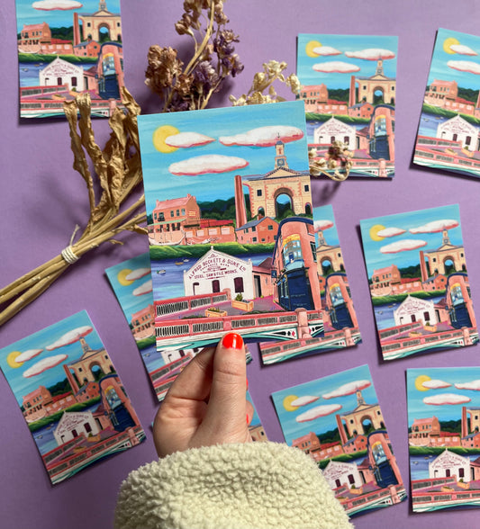 An illustration of Kelham island in Sheffield. Colourful drawings of the following places; Alfred Beckett & Sons Ltd, The Fat Cat Pub, The Riverside Pub, River Don, Green Lane Works Clocktower, The Chimney House. Set against a blue sky with fluffy clouds and the shining sunshine. An illustration with lots of blue and pinks. A small signature in white the corner. Designed by Rebecca Gibbs on a postcard at Rebecca Elizabeth Draws.