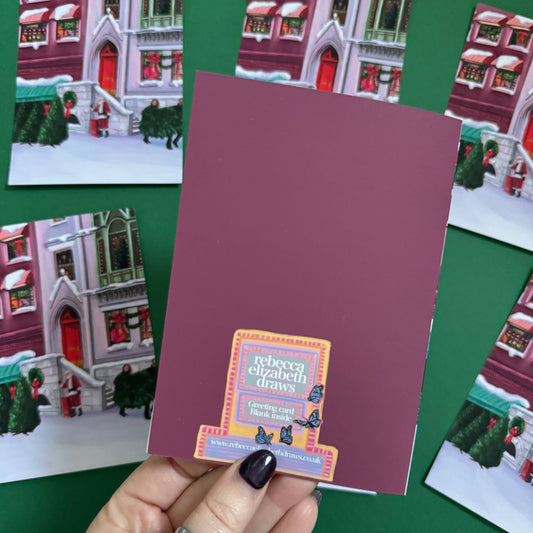 Christmas Illustration inspired by the film When Harry Met Sally. With Harry & Sally running off with their Christmas tree at the right hand side, the tree shop to the left and a background of new York buildings with Christmas décor peaking out the windows. Christmas card with red back designed by Rebecca Gibbs at Rebecca Elizabeth Draws.