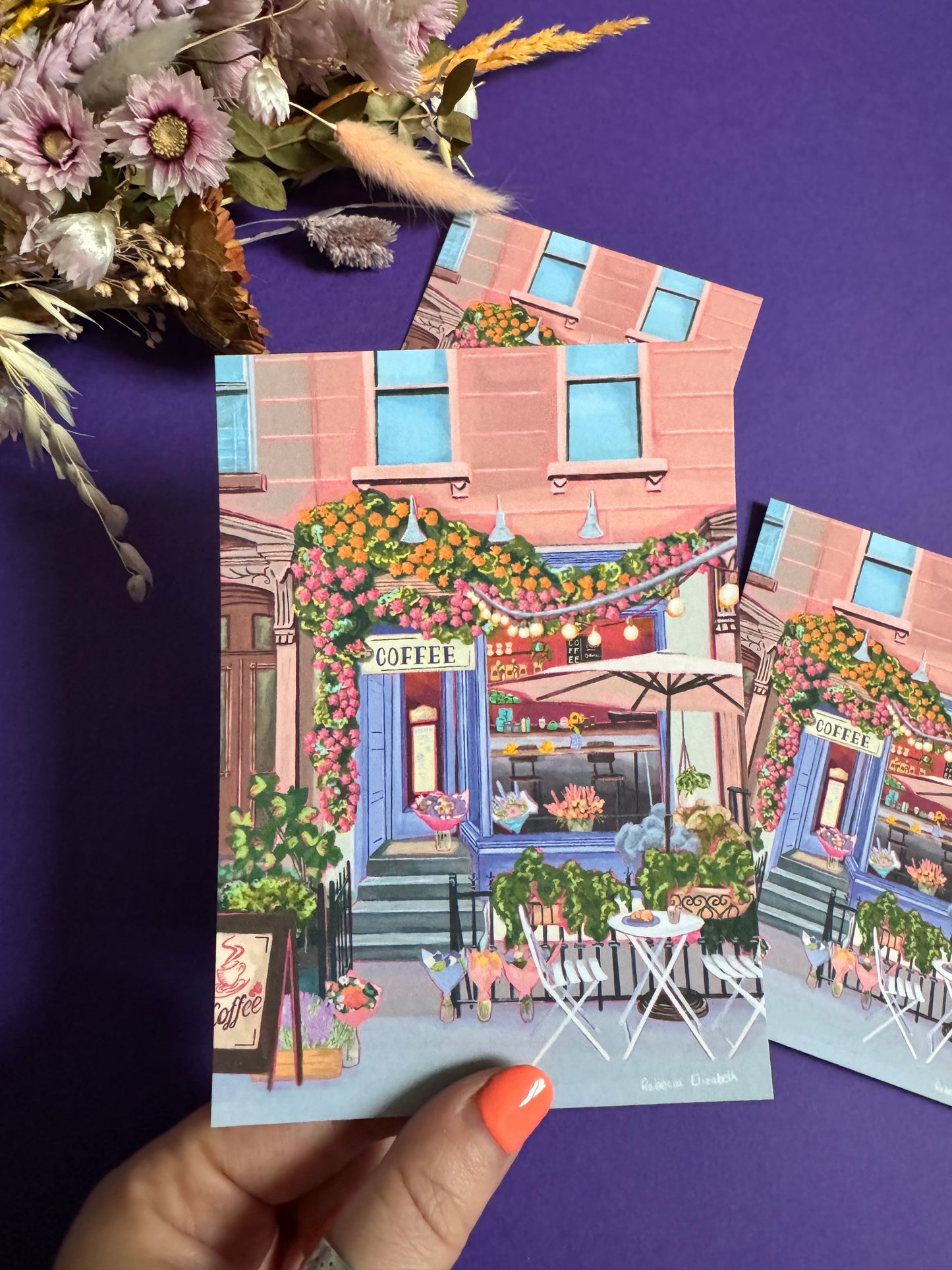 A coffee shop illustration with a blue exterior, an outside table and umbrella and building windows surrounding. This illustration is filled with lots of plants and flowers and features cups of coffee and baked goods. Illustrated by Rebecca Elizabeth Draws.