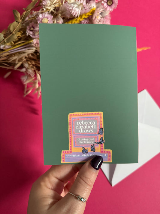 Illustration on a greeting card of a van opened up showing lots of bookshelves stacked with books, art frames out front and lots of blossoming pink, orange and blue flowers over the top. A big tree with pink roses over hangs and behind sits old fashioned buildings with quaint architecture and balconies. A whimsical fantasy print.