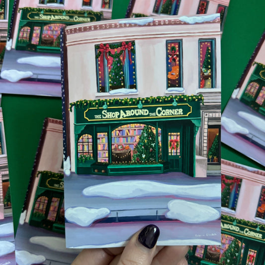 Christmas Illustration inspired by the film You’ve Got Mail. The Shop Around the Corner is a cosy & whimsical bookshop owned by Kathleen Kelly, inside is lots of fairy lights, Christmas trees, books and baubles. Outside you can see snow and inside the cosy windows with festive decor. Christmas card with green back designed by Rebecca Gibbs at Rebecca Elizabeth Draws.