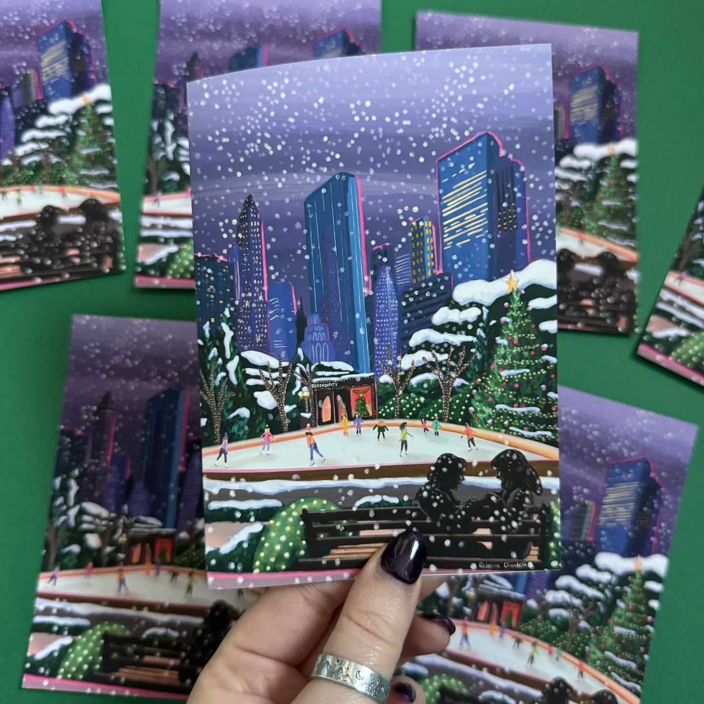 Christmas Illustration inspired by the film Serendipity. The main stars are silhouetted at the front of this illustration with the new York skyline behind them, at the base of the high rise buildings are Christmas trees, the serendipity shop and an ice skate rink with people on. A christmas card with a blue back designed by Rebecca Gibbs at Rebecca Elizabeth Draws.