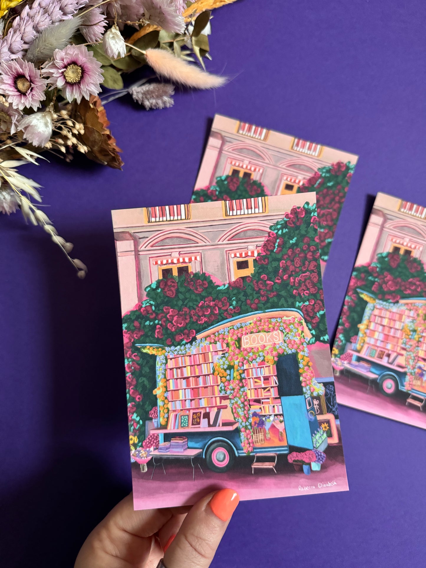 Illustration of a van opened up showing lots of bookshelves stacked with books, art frames out front and lots of blossoming pink, orange and blue flowers over the top. A big tree with pink roses over hangs and behind sits old fashioned buildings with quaint architecture and balconies. A whimsical fantasy print.