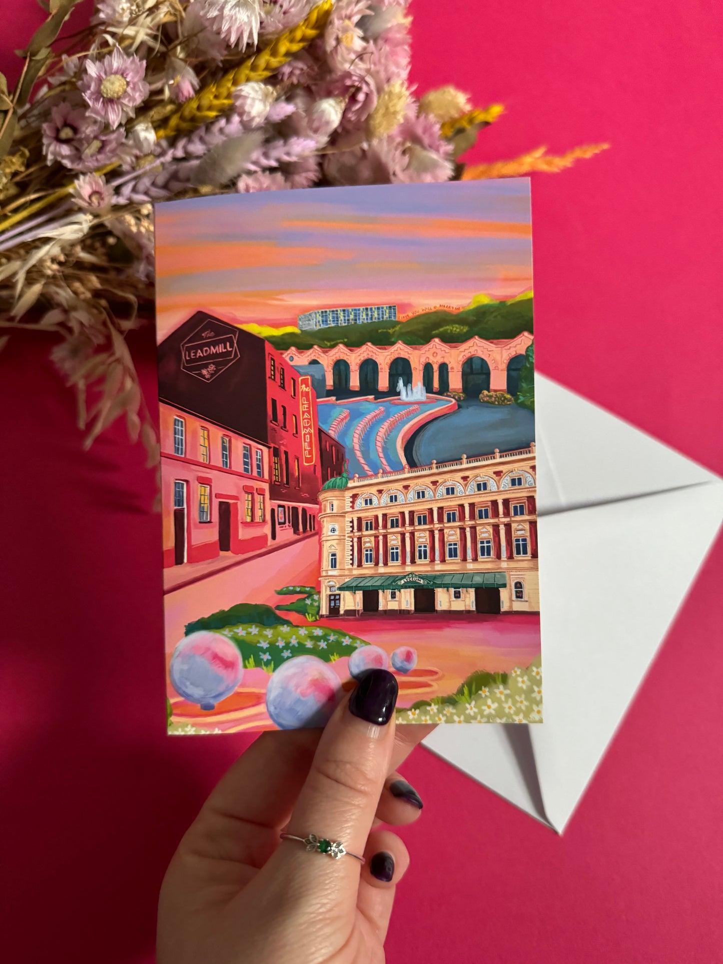 An illustrated greeting card of Sheffield city, Yorkshire. Featuring The Leadmill, the Lyceum, Sheffield train station, Sheffield Park Hill flats, park hill graffiti and water features. This illustration is very vibrant with bright pinks and oranges, the places featured are collaged together against a purple and orange sky. There is a small signature in white the corner. Designed by Rebecca Gibbs at Rebecca Elizabeth Draws.