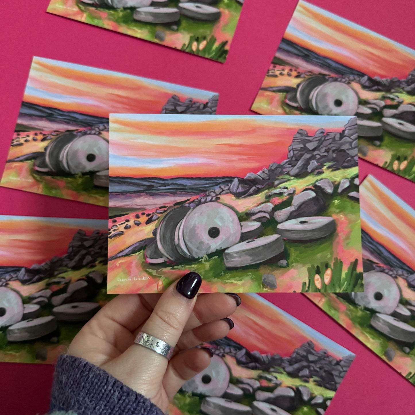 Colourful illustrated postcard of the millstones at Stanage edge in Derbyshire. With lots of pinks, greens and greys and a sunset sky. Designed by Rebecca Elizabeth Draws.