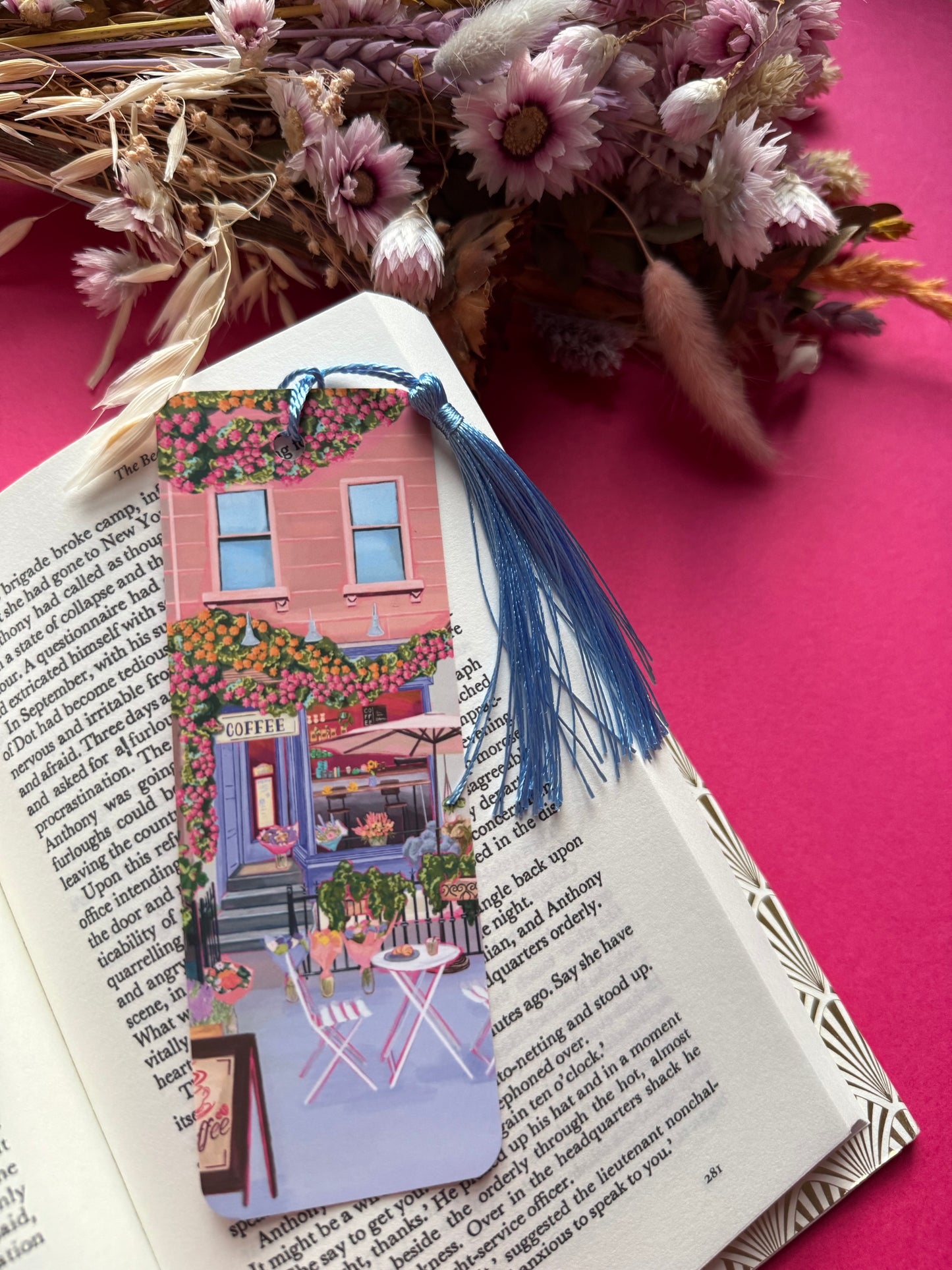 A coffee shop illustrated bookmark with a blue exterior, an outside table and umbrella and building windows surrounding. This illustration is filled with lots of plants and flowers and features cups of coffee and baked goods. Illustrated by Rebecca Elizabeth Draws.