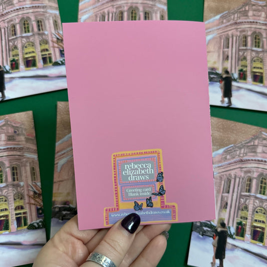 Christmas Illustration inspired by the film Bridget Jones Diary. The end scene featuring Mark Darcy & Bridget Jones kissing in the snow on a hazy street in London in front of the Montblanc diary shop. In the background you can see the mini her friends drive away in and a poster with Marks reindeer Christmas jumper. A christmas card with a pink back designed by Rebecca Gibbs at Rebecca Elizabeth Draws.