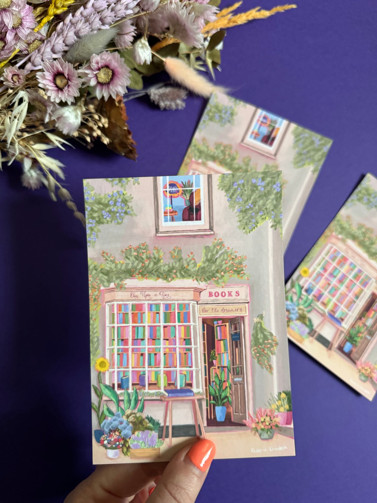 An illustration of a cute bookshop, the window shows the bookshelves inside full of bright coloured book spines. There are books placed stood up at the front. Outside the building is a soft colour with lots of greenery in soft pale greens and orange and lilac flowers. In front of the building there are plant pots and flowers. There is a small signature in white the corner. Designed by Rebecca Gibbs on a postcard at Rebecca Elizabeth Draws.