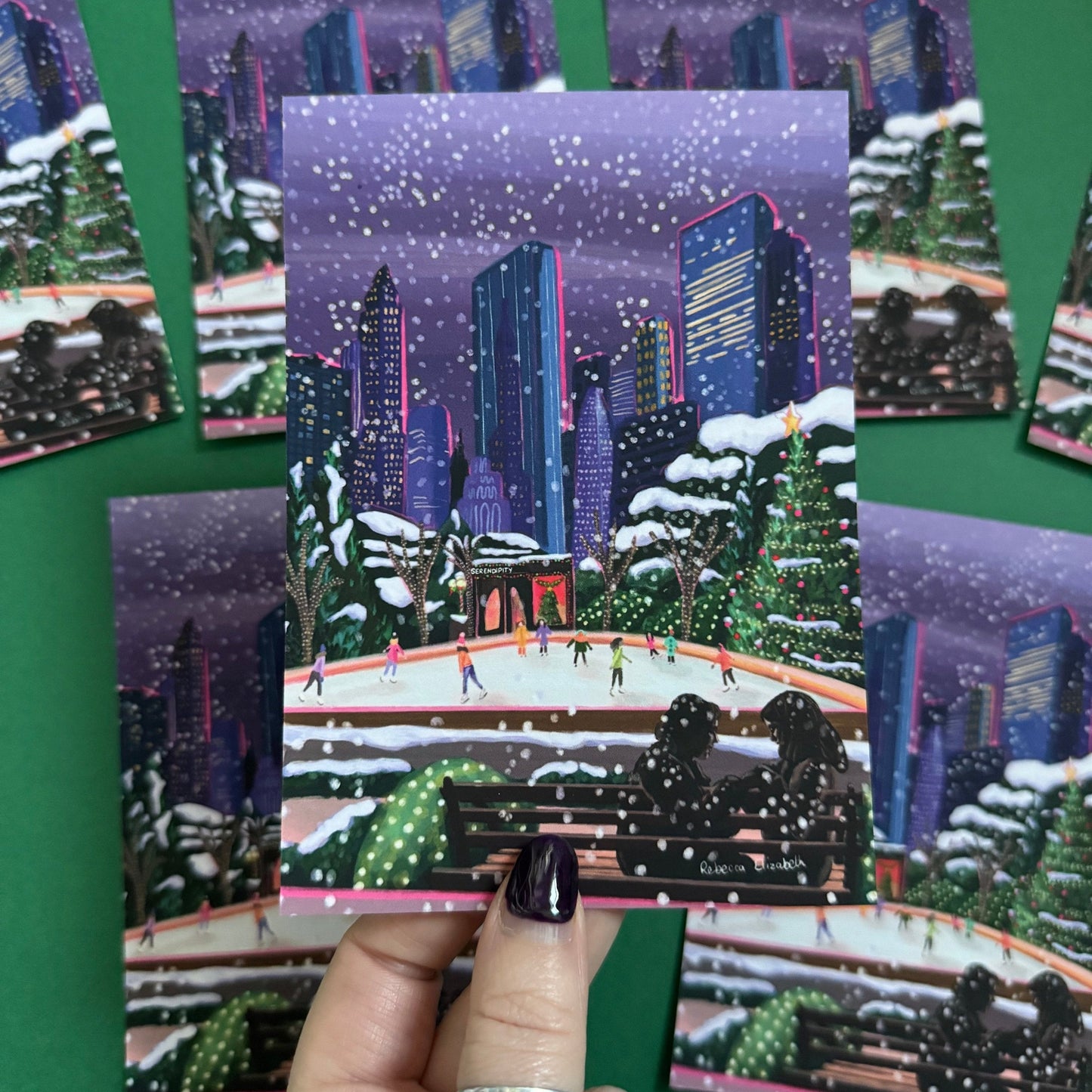 Christmas Illustrated postcard inspired by the film Serendipity. The main stars are silhouetted at the front of this illustration with the new York skyline behind them, at the base of the high rise buildings are Christmas trees, the serendipity shop and an ice skate rink with people on. Designed by Rebecca Gibbs at Rebecca Elizabeth Draws.