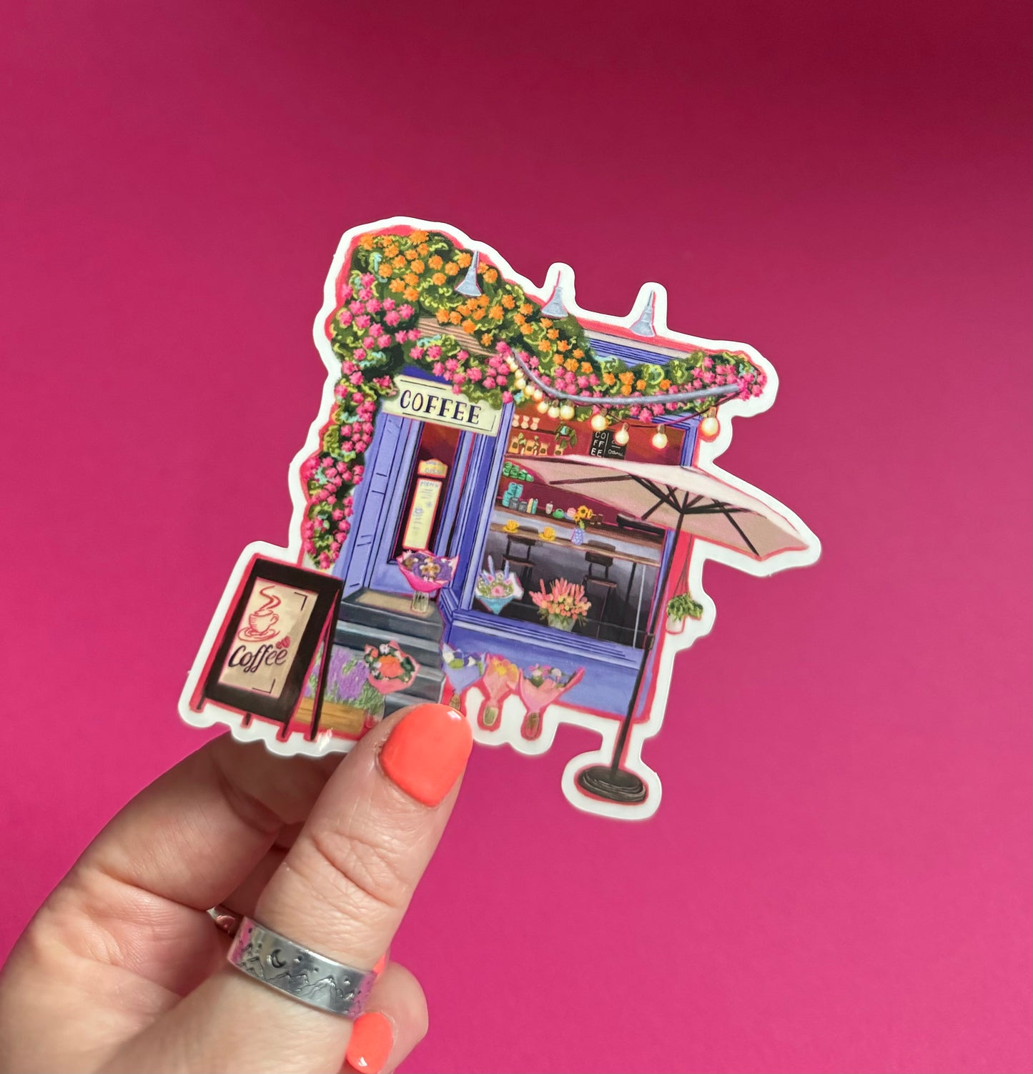 A coffee shop art sticker with a blue exterior, an outside table and umbrella and building windows surrounding. This sticker is filled with lots of plants and flowers & features cups of coffee & baked goods. Illustrated by Rebecca Elizabeth Draws.