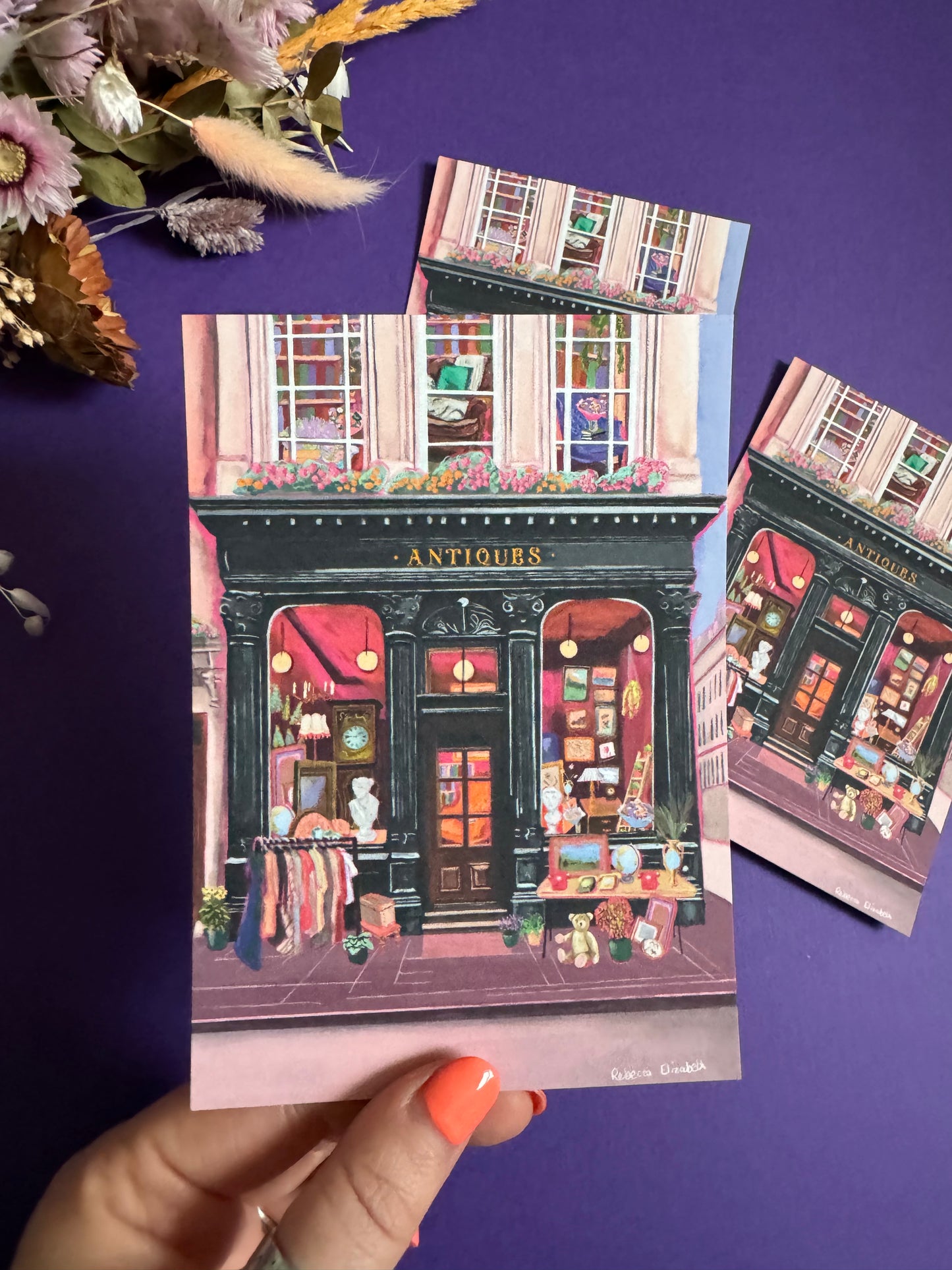 Illustration of a antiques shop front in black with ornate designs, inside is pink and golden with vintage objects such a clocks, statues, picture frames. The illustration gives off dark academia vibes. Designed by Rebecca Elizabeth Draws.
