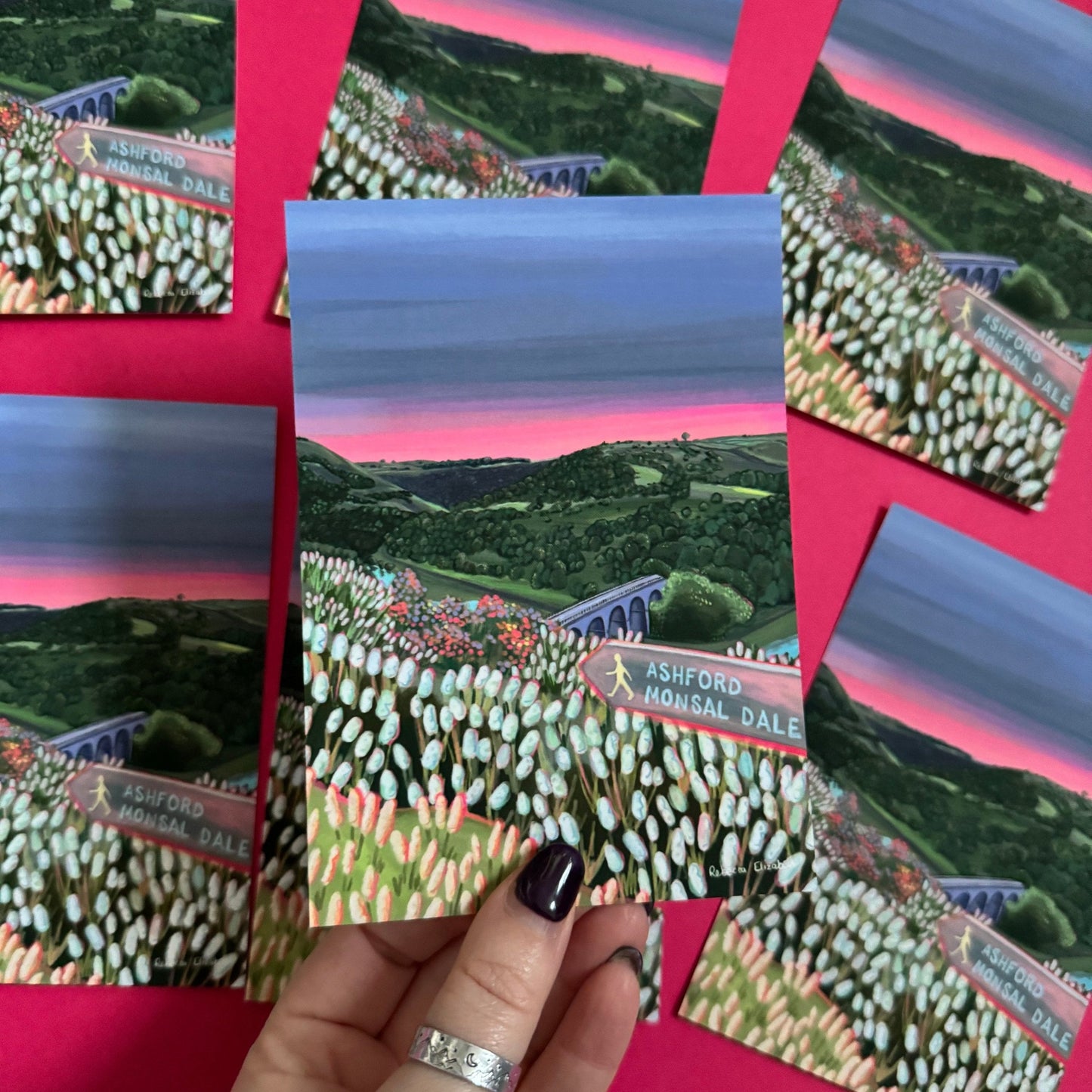 Postcard art of Monsal Head in Derbyshire, Peak District. Rolling green hills filled with trees, an old viaduct over the river Wye and white flowers in the foreground. The sky is a setting sun with pink on the horizon fading into dark blue grey.