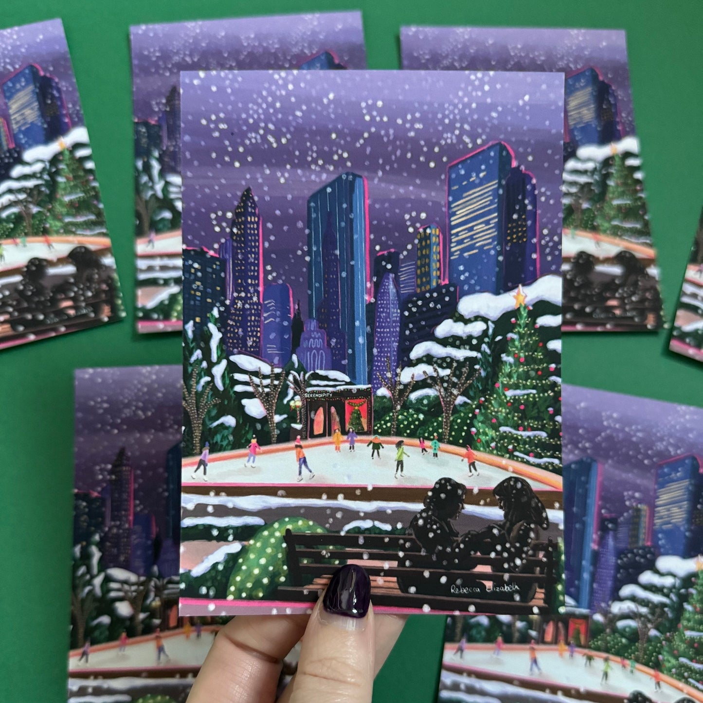 Christmas Illustrated postcard inspired by the film Serendipity. The main stars are silhouetted at the front of this illustration with the new York skyline behind them, at the base of the high rise buildings are Christmas trees, the serendipity shop and an ice skate rink with people on. Designed by Rebecca Gibbs at Rebecca Elizabeth Draws.