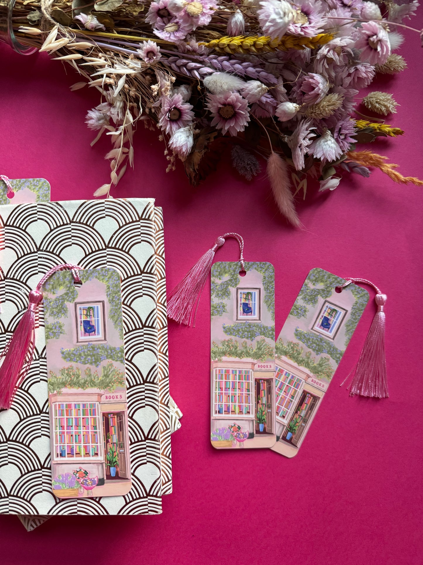 An illustrated bookmark of a cute cosy bookshop, the window shows the bookshelves inside full of bright coloured book spines. There are books placed stood up at the front. Outside the building is a soft colour with lots of greenery in soft pale greens and orange and lilac flowers. In front of the building there are plant pots and flowers. A whimsical cosy art bookmark. There is a small signature in white the corner. Designed by Rebecca Gibbs at Rebecca Elizabeth Draws.