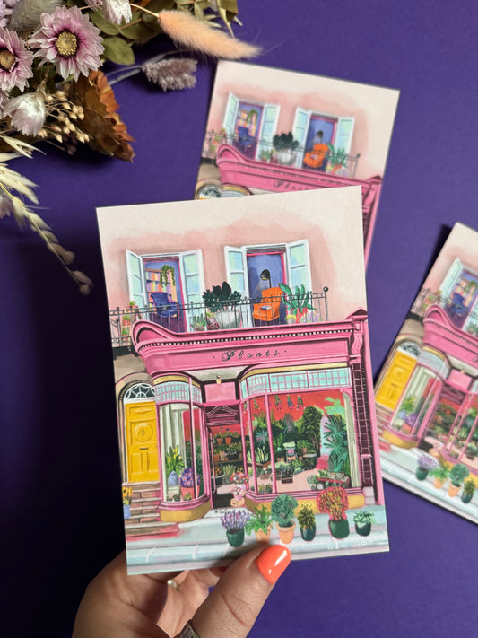 Pink shop front of a plant store with lots of greenery inside, a balcony above with windows showing chairs and a bookcase and more flowers. A yellow door to the side with a sunflower vase resting on the steps by Rebecca Elizabeth Draws.