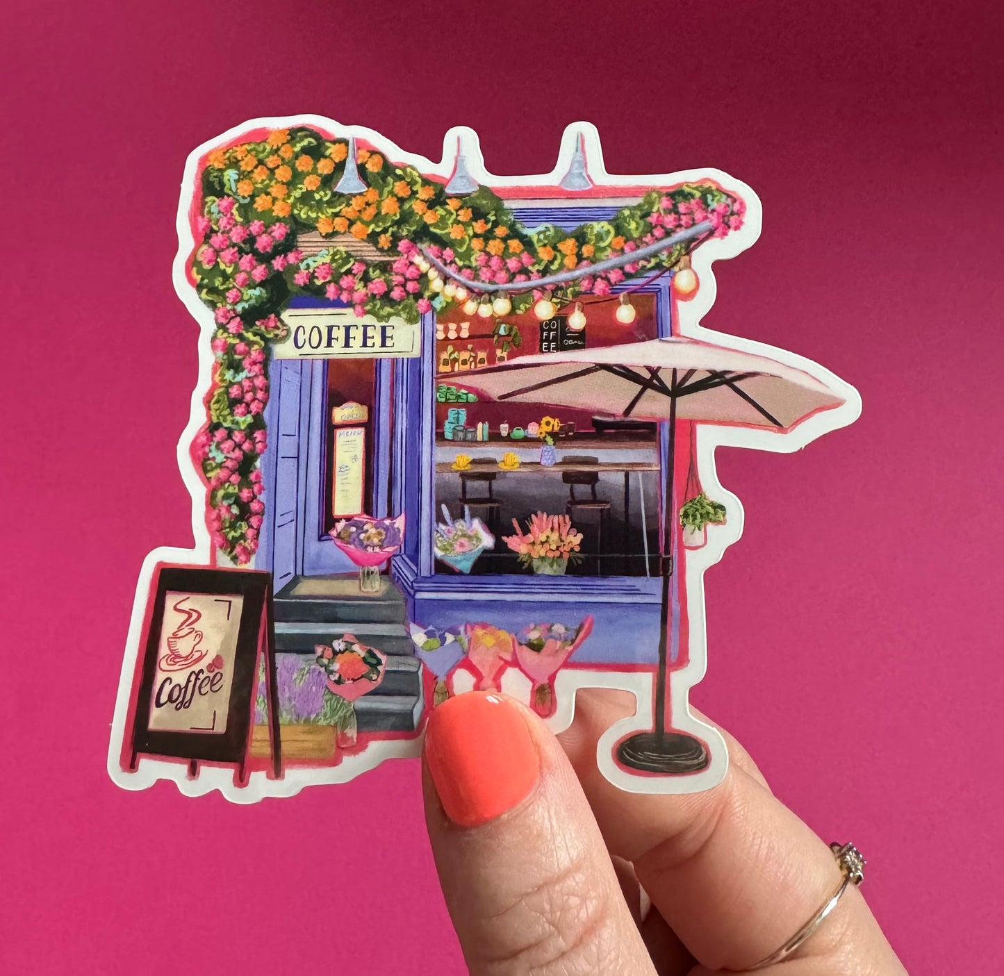 A coffee shop art sticker with a blue exterior, an outside table and umbrella and building windows surrounding. This sticker is filled with lots of plants and flowers & features cups of coffee & baked goods. Illustrated by Rebecca Elizabeth Draws.