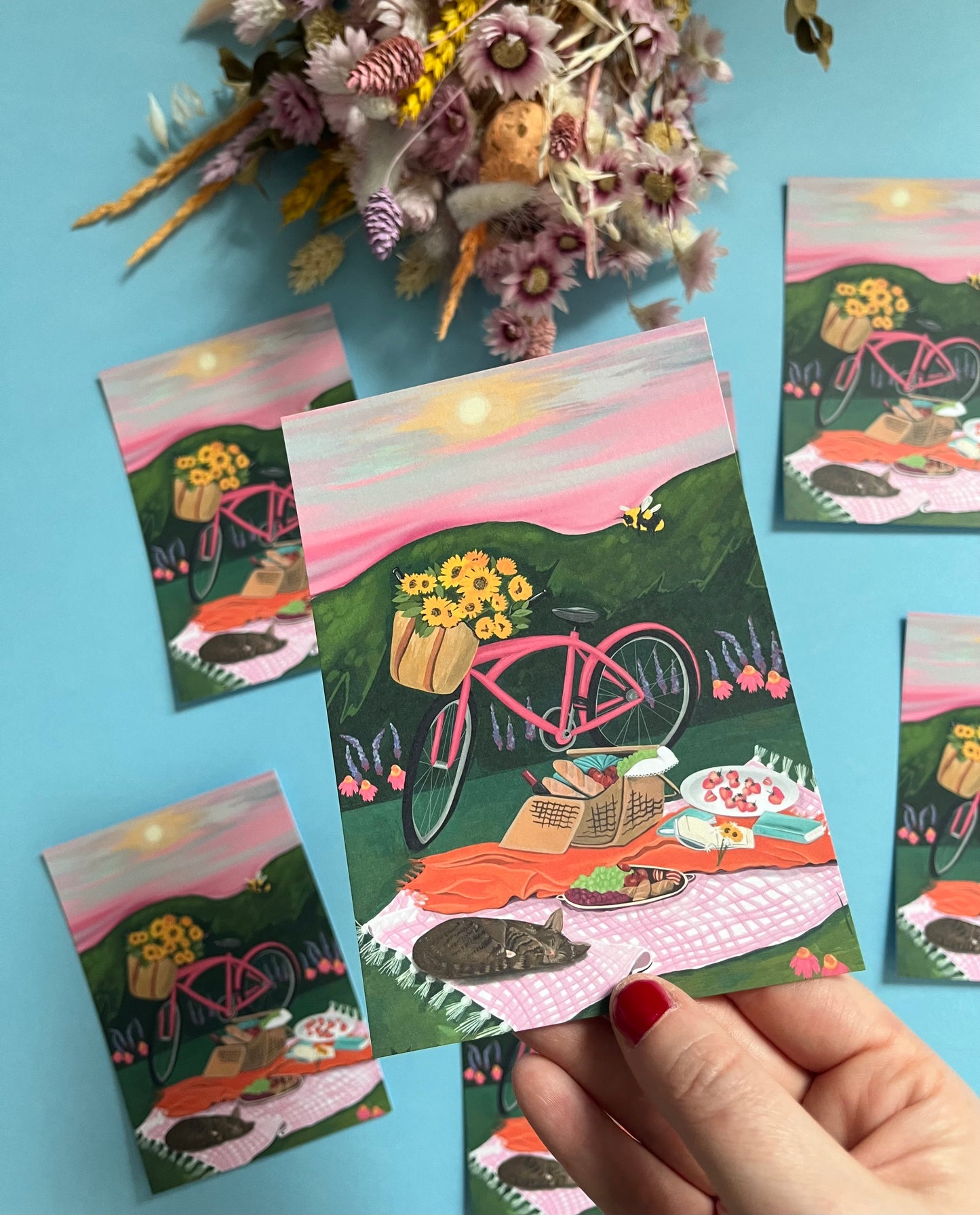 An illustration of a pink bicycle resting up by a picnic blanket filled with wine, loaves of bread, strawberries, picked sunflowers and a book. There is a cat sleeping and behind is pink and purple flowers against the greenery. There is a bumble bee and the sky is pink and grey with a small yellow sun. There is a small signature in white the corner. Designed by Rebecca Gibbs on a postcard at Rebecca Elizabeth Draws.