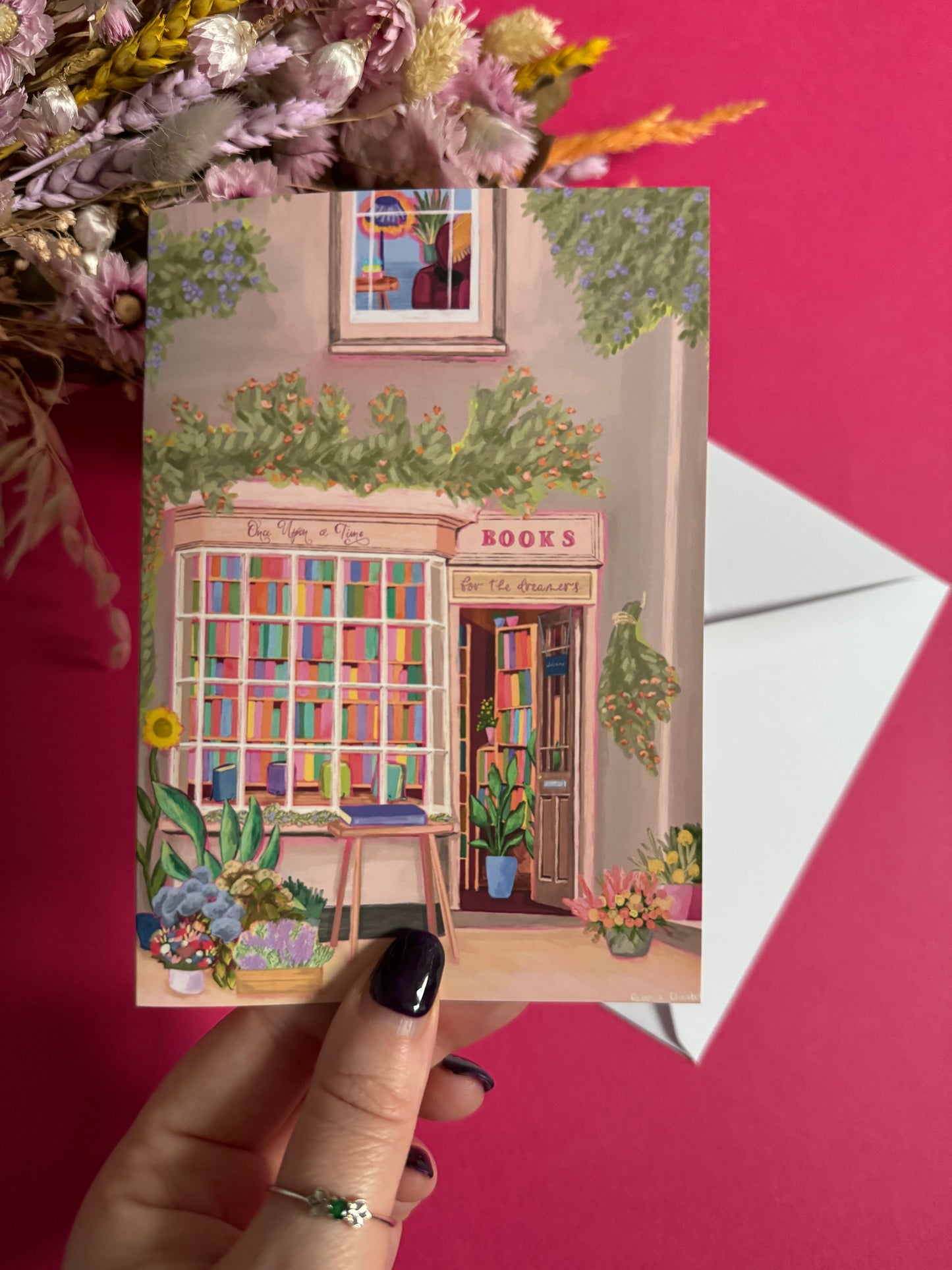 An illustration of a cute whimsical bookshop, the window shows the bookshelves inside full of bright coloured book spines with lots of flowers. The back of the card is pink with the Rebecca Elizabeth Draws logo.
