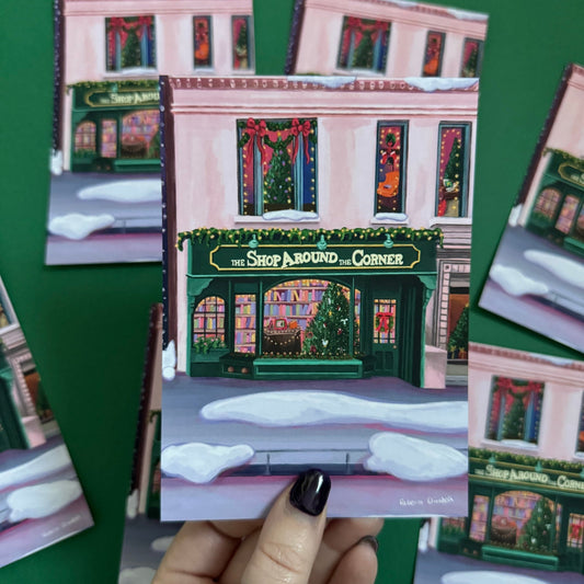 Christmas Illustrated postcard inspired by the film You’ve Got Mail. The Shop Around the Corner is a cosy & whimsical bookshop owned by Kathleen Kelly, inside is lots of fairy lights, Christmas trees, books and baubles. Outside you can see snow and inside the cosy windows with festive decor. Designed by Rebecca Gibbs at Rebecca Elizabeth Draws.