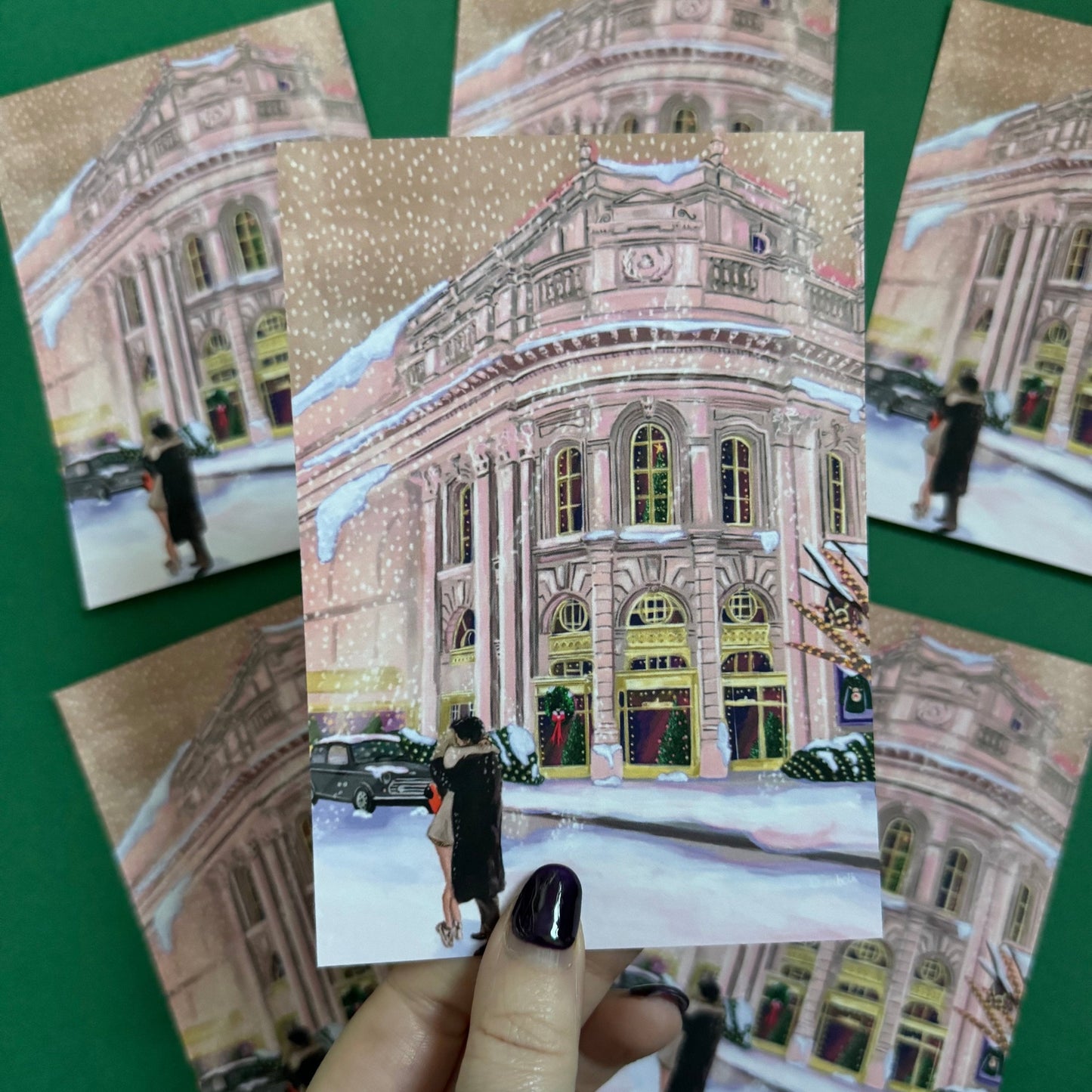 Christmas Illustrated postcard inspired by the film Bridget Jones Diary. The end scene featuring Mark Darcy & Bridget Jones kissing in the snow on a hazy street in London in front of the Montblanc diary shop. In the background you can see the mini her friends drive away in and a poster with Marks reindeer Christmas jumper. A velvet laminated postcard. Designed by Rebecca Gibbs at Rebecca Elizabeth Draws.