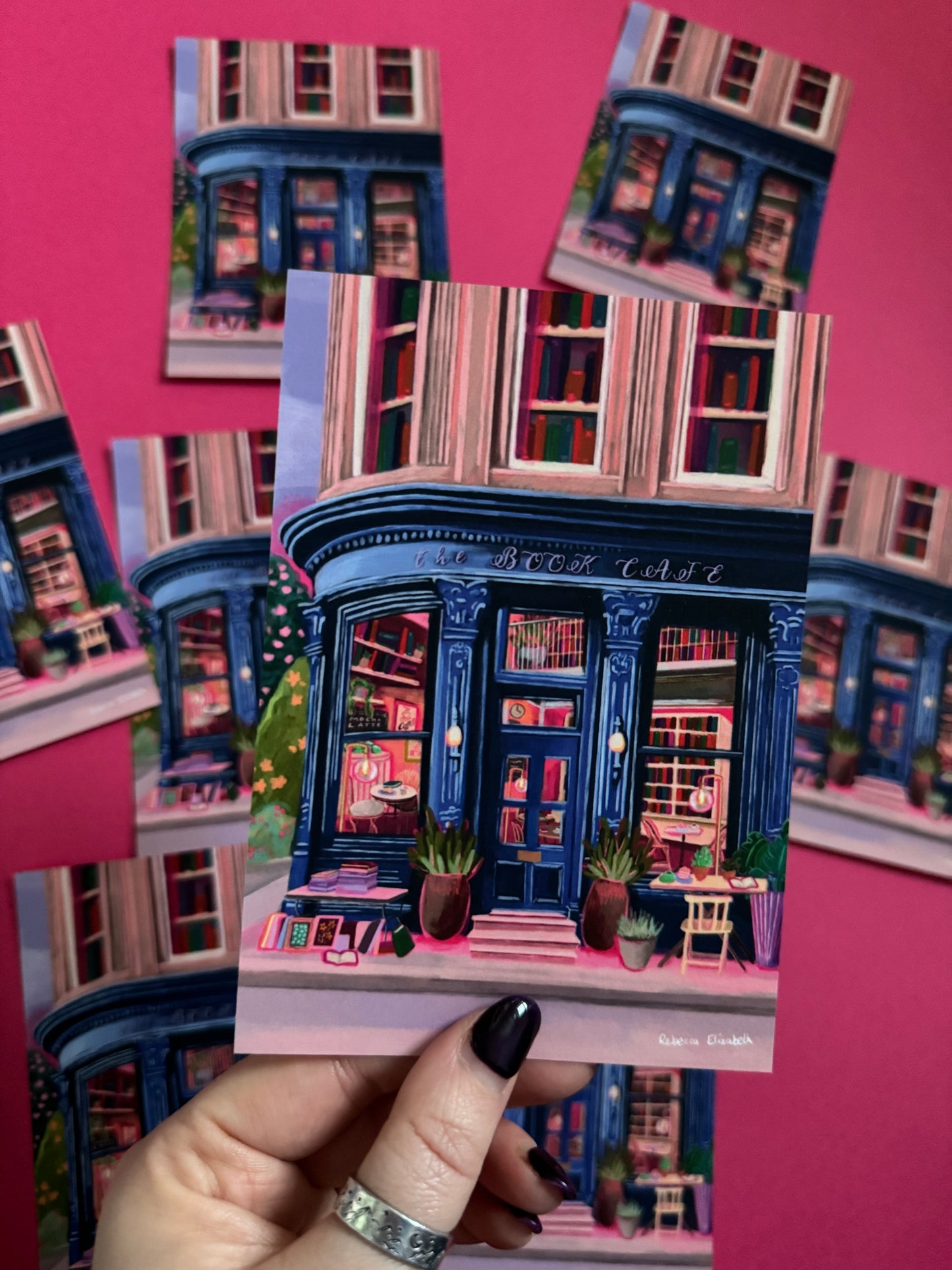 Store front illustration in a deep blue with antique vintage design, inside the book café is a bookcase with jewel tone books, a coffee stand with mugs & cake. The are tables inside and out and upstairs are 3 windows with shelves of books inside. Designed by Rebecca Elizabeth Draws.
