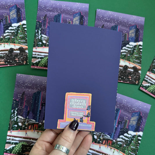 Christmas Illustration inspired by the film Serendipity. The main stars are silhouetted at the front of this illustration with the new York skyline behind them, at the base of the high rise buildings are Christmas trees, the serendipity shop and an ice skate rink with people on. A christmas card with a blue back designed by Rebecca Gibbs at Rebecca Elizabeth Draws.