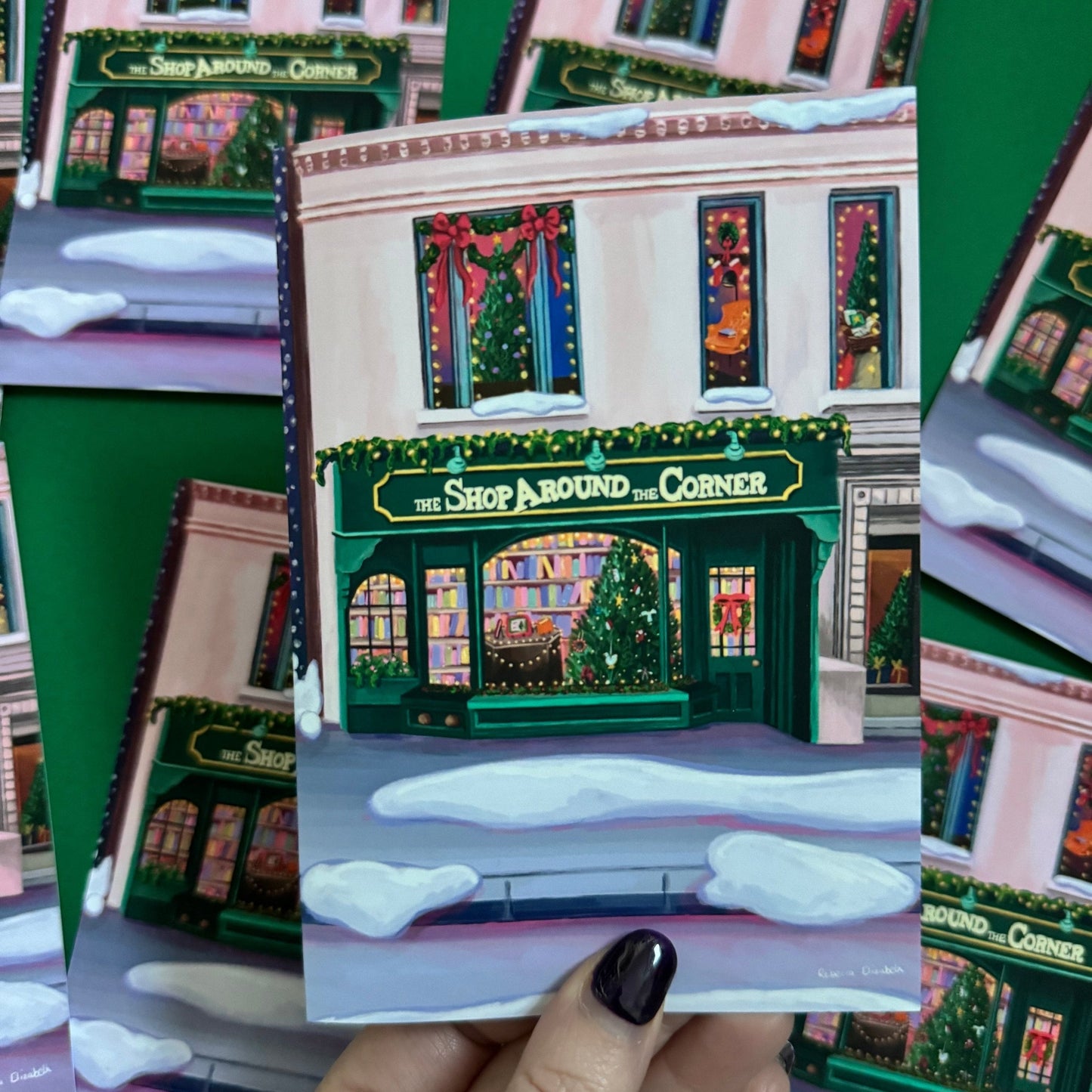 Christmas Illustration inspired by the film You’ve Got Mail. The Shop Around thChristmas Illustration inspired by the film You’ve Got Mail. The Shop Around the Corner is a cosy & whimsical bookshop owned by Kathleen Kellye Corner is a cosy & whimsical bookshop owned by Kathleen Kelly, inside is lots of fairy lights, Christmas trees, books and baubles. Outside you can see snow and inside the cosy windows with festive decor. Christmas card with green back designed by Rebecca Gibbs at Rebecca Elizabeth Draws.