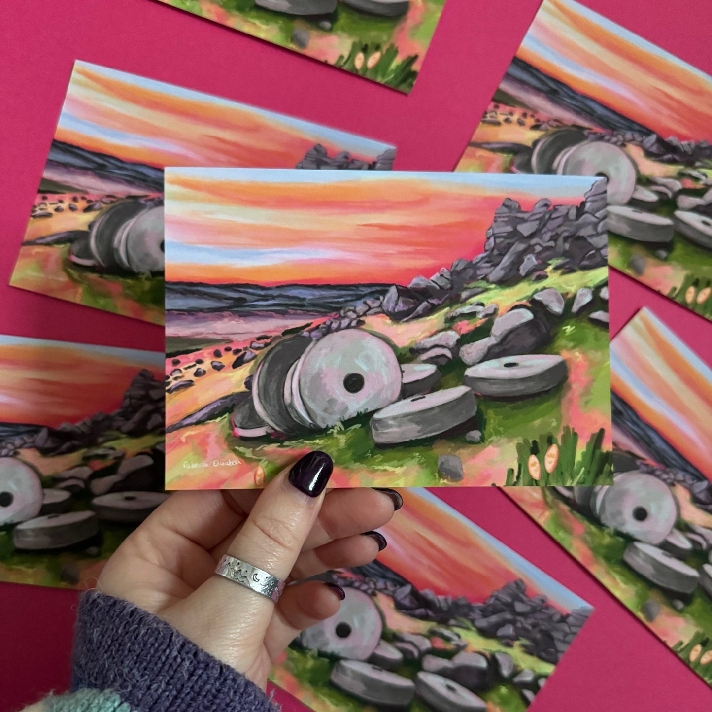 Colourful illustrated postcard of the millstones at Stanage edge in Derbyshire. With lots of pinks, greens and greys and a sunset sky. Designed by Rebecca Elizabeth Draws.