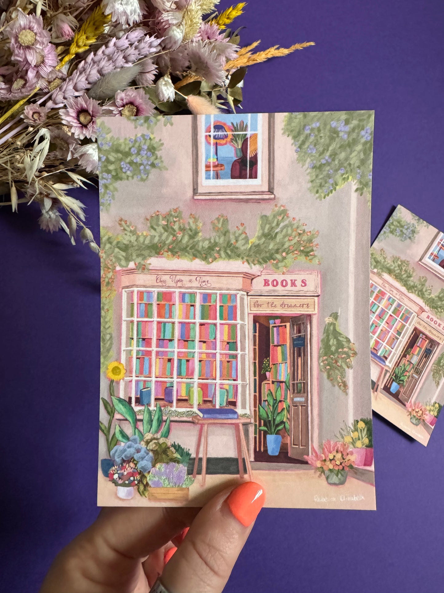 An illustration of a cute bookshop, the window shows the bookshelves inside full of bright coloured book spines. There are books placed stood up at the front. Outside the building is a soft colour with lots of greenery in soft pale greens and orange and lilac flowers. In front of the building there are plant pots and flowers. There is a small signature in white the corner. Designed by Rebecca Gibbs on a postcard at Rebecca Elizabeth Draws.