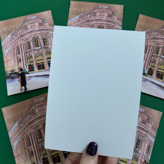 Christmas Illustrated postcard inspired by the film Bridget Jones Diary. The end scene featuring Mark Darcy & Bridget Jones kissing in the snow on a hazy street in London in front of the Montblanc diary shop. In the background you can see the mini her friends drive away in and a poster with Marks reindeer Christmas jumper. A velvet laminated postcard. Designed by Rebecca Gibbs at Rebecca Elizabeth Draws.