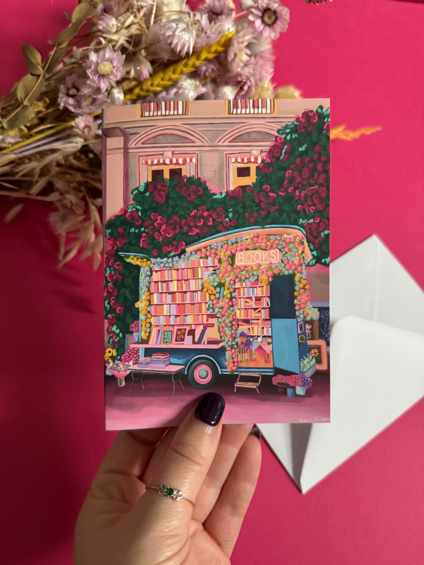 Illustration on a greeting card of a van opened up showing lots of bookshelves stacked with books, art frames out front and lots of blossoming pink, orange and blue flowers over the top. A big tree with pink roses over hangs and behind sits old fashioned buildings with quaint architecture and balconies. A whimsical fantasy print.