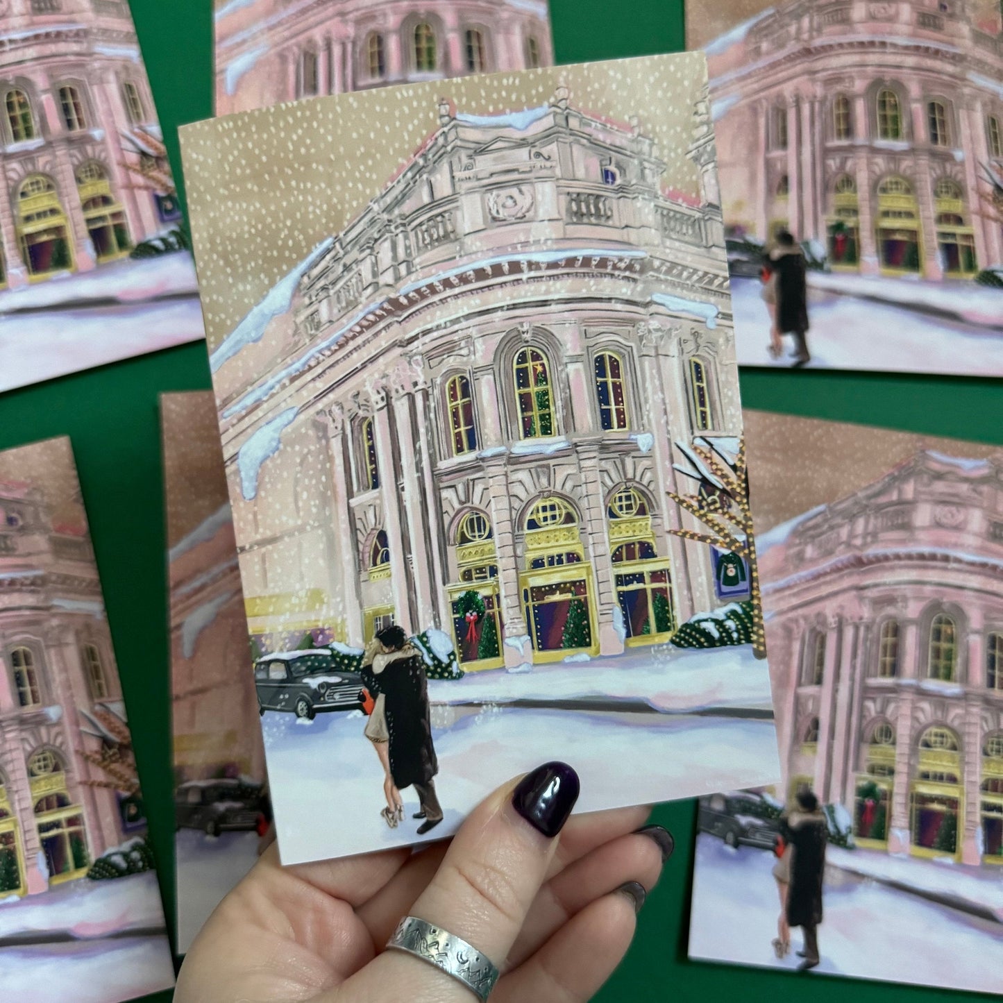 Christmas Illustration inspired by the film Bridget Jones Diary. The end scene featuring Mark Darcy & Bridget Jones kissing in the snow on a hazy street in London in front of the Montblanc diary shop. In the background you can see the mini her friends drive away in and a poster with Marks reindeer Christmas jumper. A christmas card with a pink back designed by Rebecca Gibbs at Rebecca Elizabeth Draws.