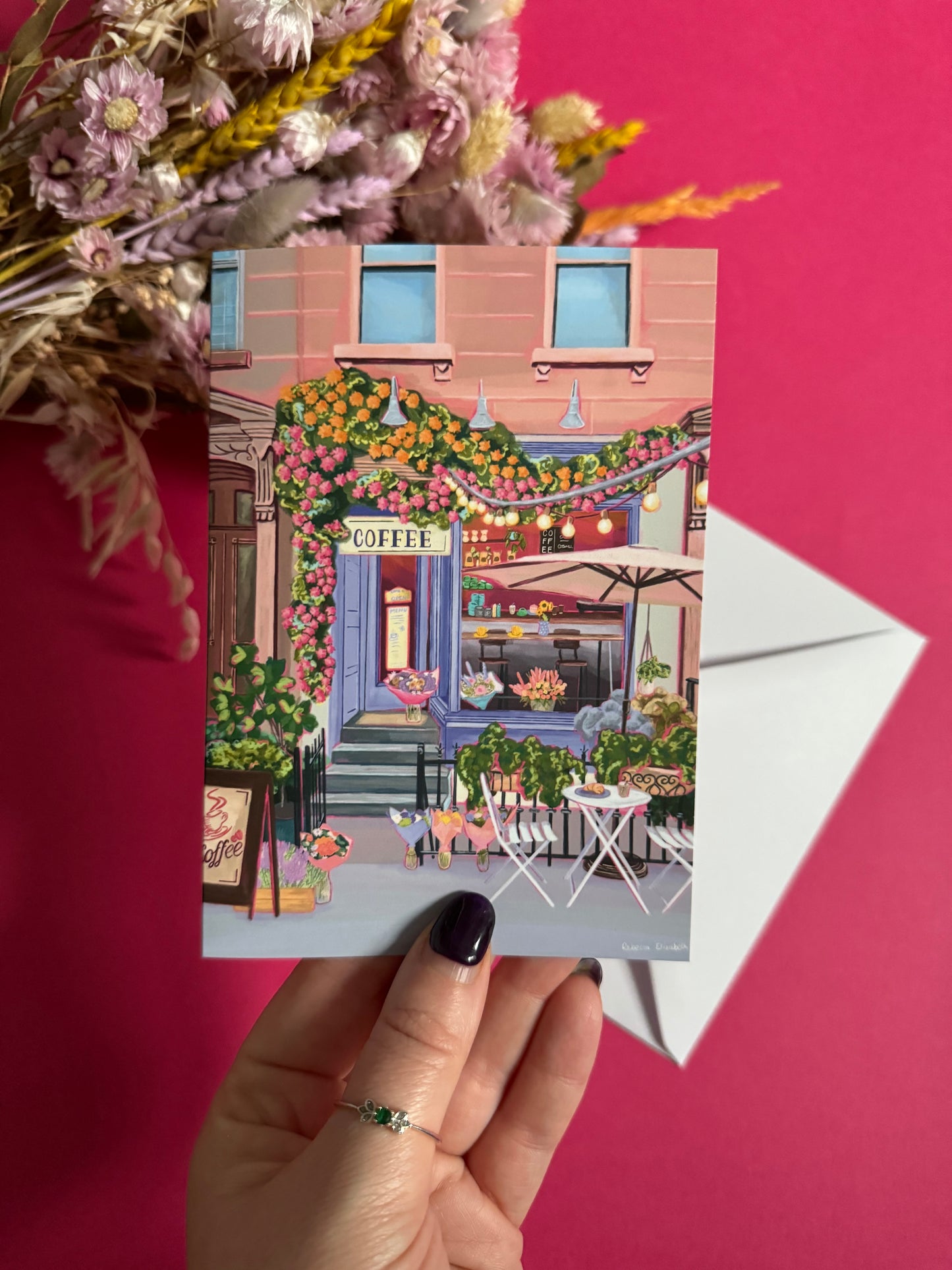 A coffee shop illustrated card with a blue exterior, an outside table and umbrella and building windows surrounding. This illustration is filled with lots of plants and flowers and features cups of coffee and baked goods. Illustrated by Rebecca Elizabeth Draws.