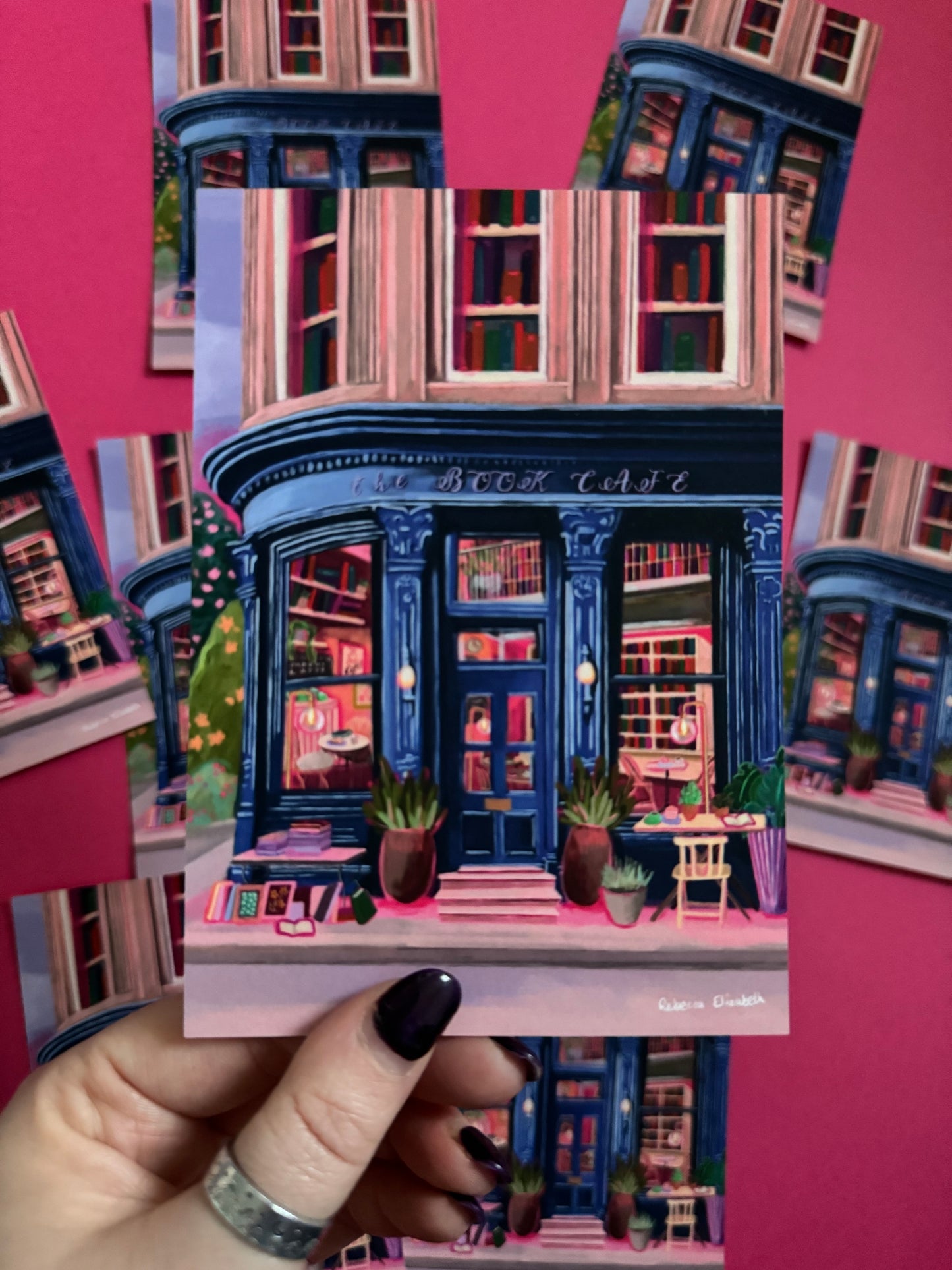 Store front illustration in a deep blue with antique vintage design, inside the book café is a bookcase with jewel tone books, a coffee stand with mugs & cake. The are tables inside and out and upstairs are 3 windows with shelves of books inside. Designed by Rebecca Elizabeth Draws.