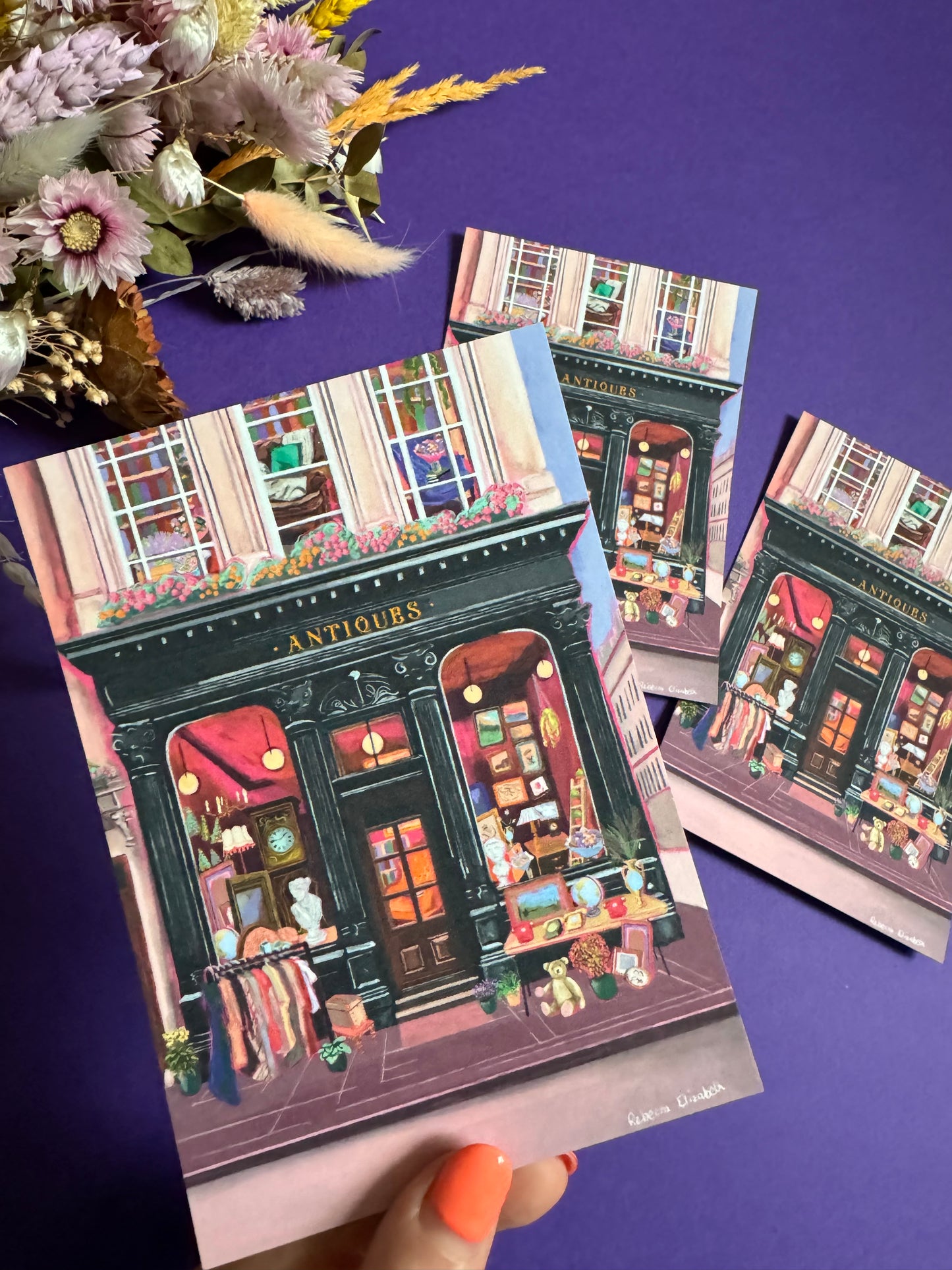 Illustration of a antiques shop front in black with ornate designs, inside is pink and golden with vintage objects such a clocks, statues, picture frames. The illustration gives off dark academia vibes. Designed by Rebecca Elizabeth Draws.