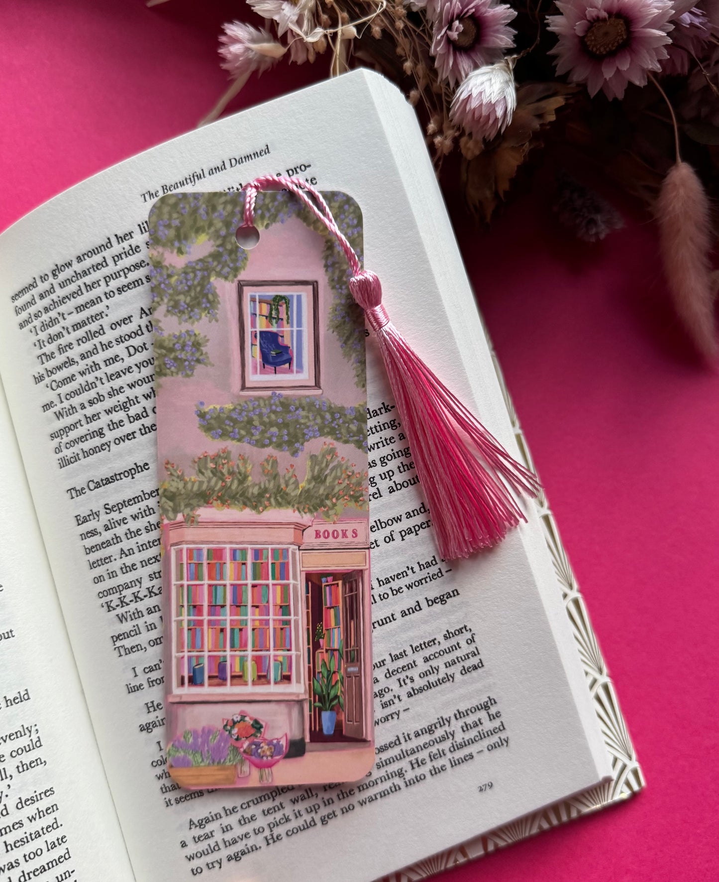An illustrated bookmark of a cute cosy bookshop, the window shows the bookshelves inside full of bright coloured book spines. There are books placed stood up at the front. Outside the building is a soft colour with lots of greenery in soft pale greens and orange and lilac flowers. In front of the building there are plant pots and flowers. A whimsical cosy art bookmark. There is a small signature in white the corner. Designed by Rebecca Gibbs at Rebecca Elizabeth Draws.