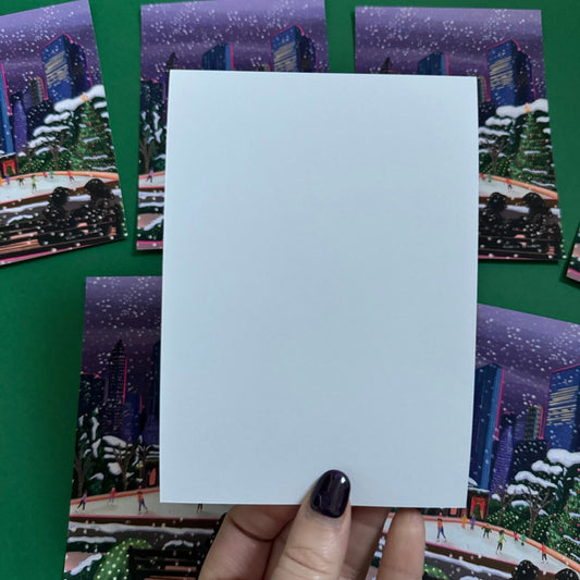 Christmas Illustrated postcard inspired by the film Serendipity. The main stars are silhouetted at the front of this illustration with the new York skyline behind them, at the base of the high rise buildings are Christmas trees, the serendipity shop and an ice skate rink with people on. Designed by Rebecca Gibbs at Rebecca Elizabeth Draws.