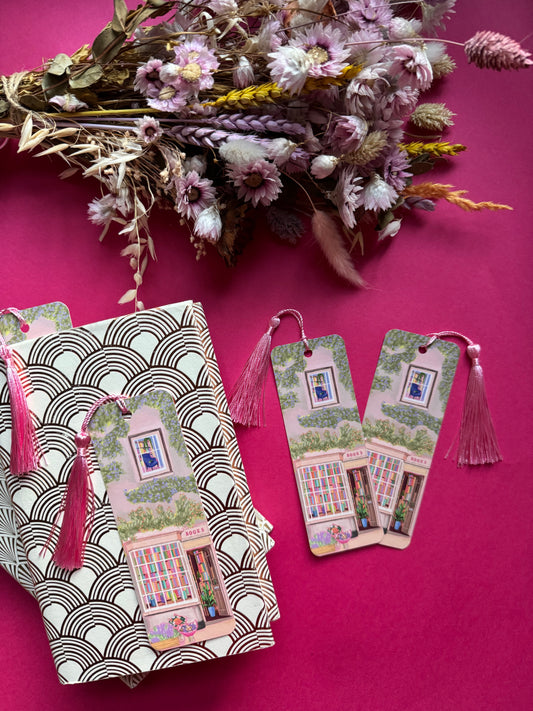 An illustrated bookmark of a cute cosy bookshop, the window shows the bookshelves inside full of bright coloured book spines. There are books placed stood up at the front. Outside the building is a soft colour with lots of greenery in soft pale greens and orange and lilac flowers. In front of the building there are plant pots and flowers. A whimsical cosy art bookmark. There is a small signature in white the corner. Designed by Rebecca Gibbs at Rebecca Elizabeth Draws.