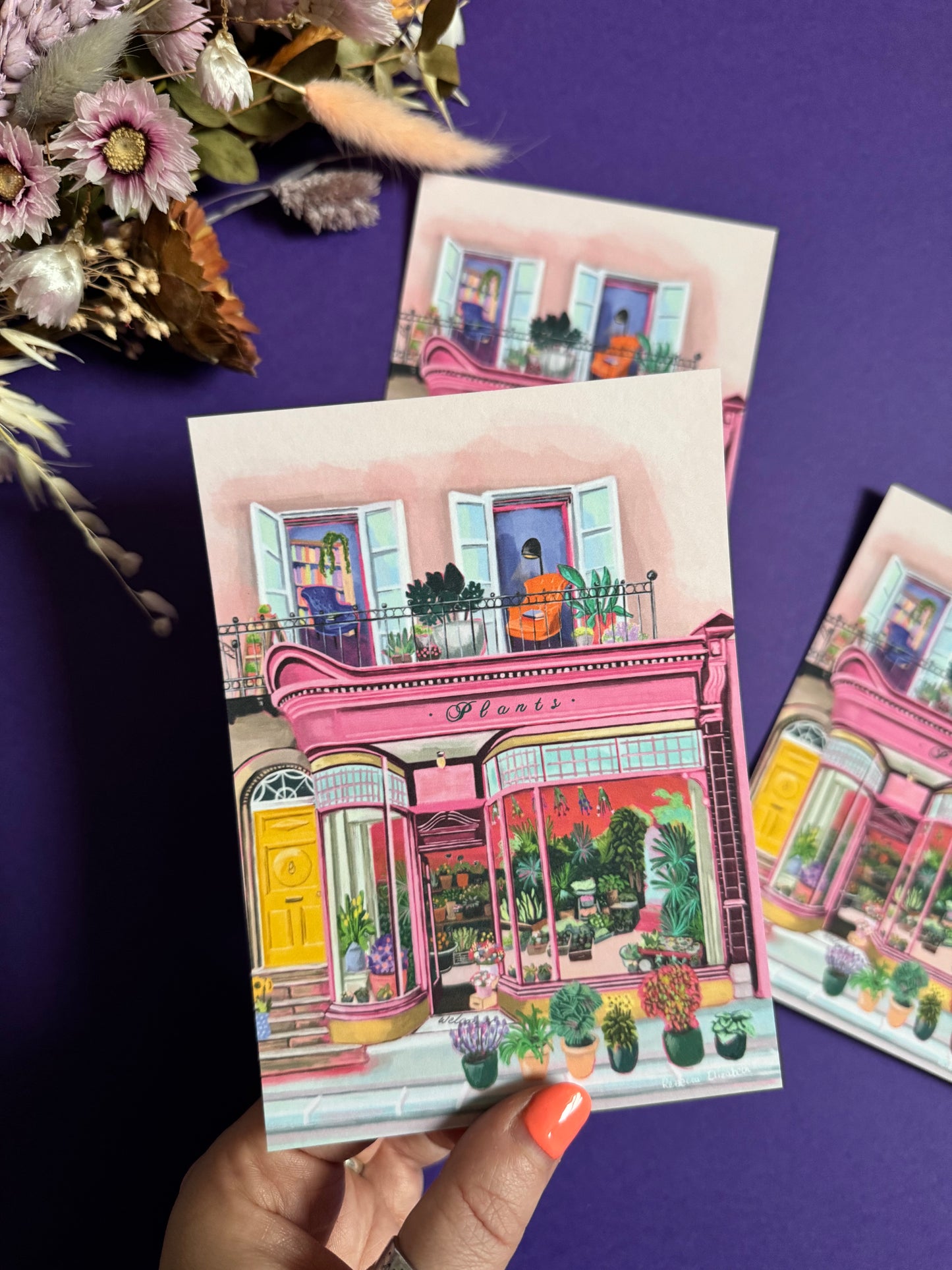 Pink shop front of a plant store with lots of greenery inside, a balcony above with windows showing chairs and a bookcase and more flowers. A yellow door to the side with a sunflower vase resting on the steps by Rebecca Elizabeth Draws.