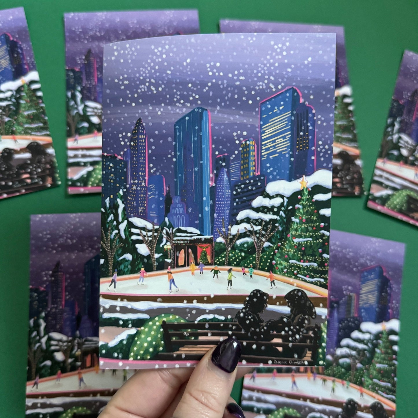 Christmas Illustration inspired by the film Serendipity. The main stars are silhouetted at the front of this illustration with the new York skyline behind them, at the base of the high rise buildings are Christmas trees, the serendipity shop and an ice skate rink with people on. A christmas card with a blue back designed by Rebecca Gibbs at Rebecca Elizabeth Draws.