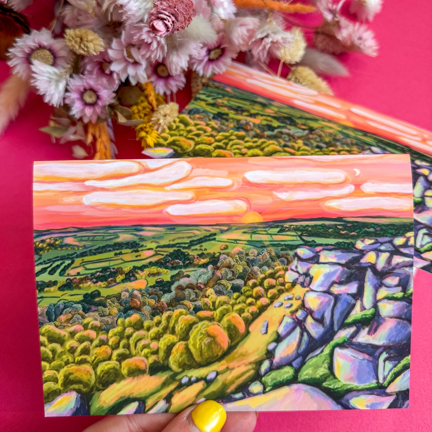 Colorful landscape painting held against a pink background with flowers.