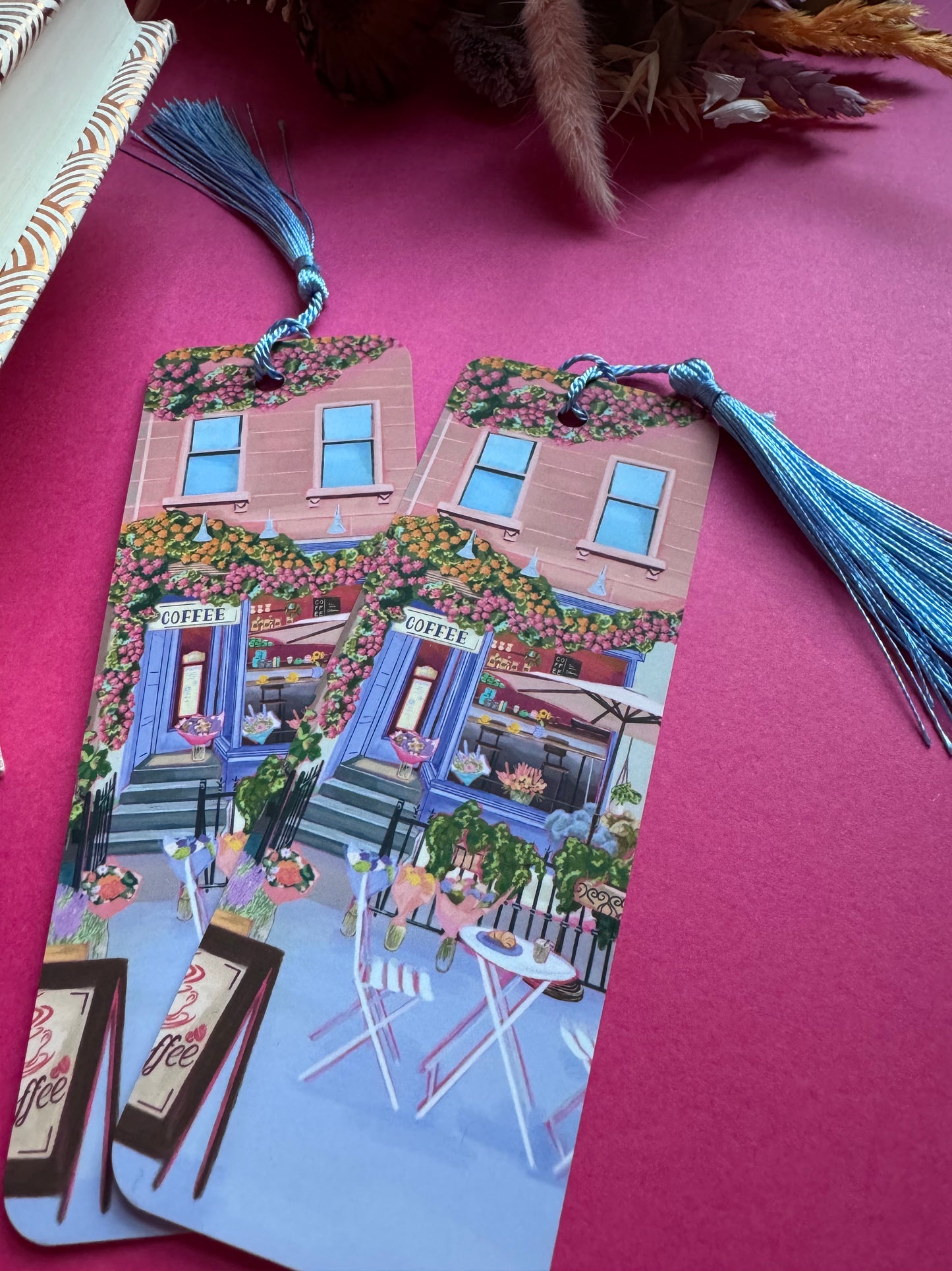 A coffee shop illustrated bookmark with a blue exterior, an outside table and umbrella and building windows surrounding. This illustration is filled with lots of plants and flowers and features cups of coffee and baked goods. Illustrated by Rebecca Elizabeth Draws.