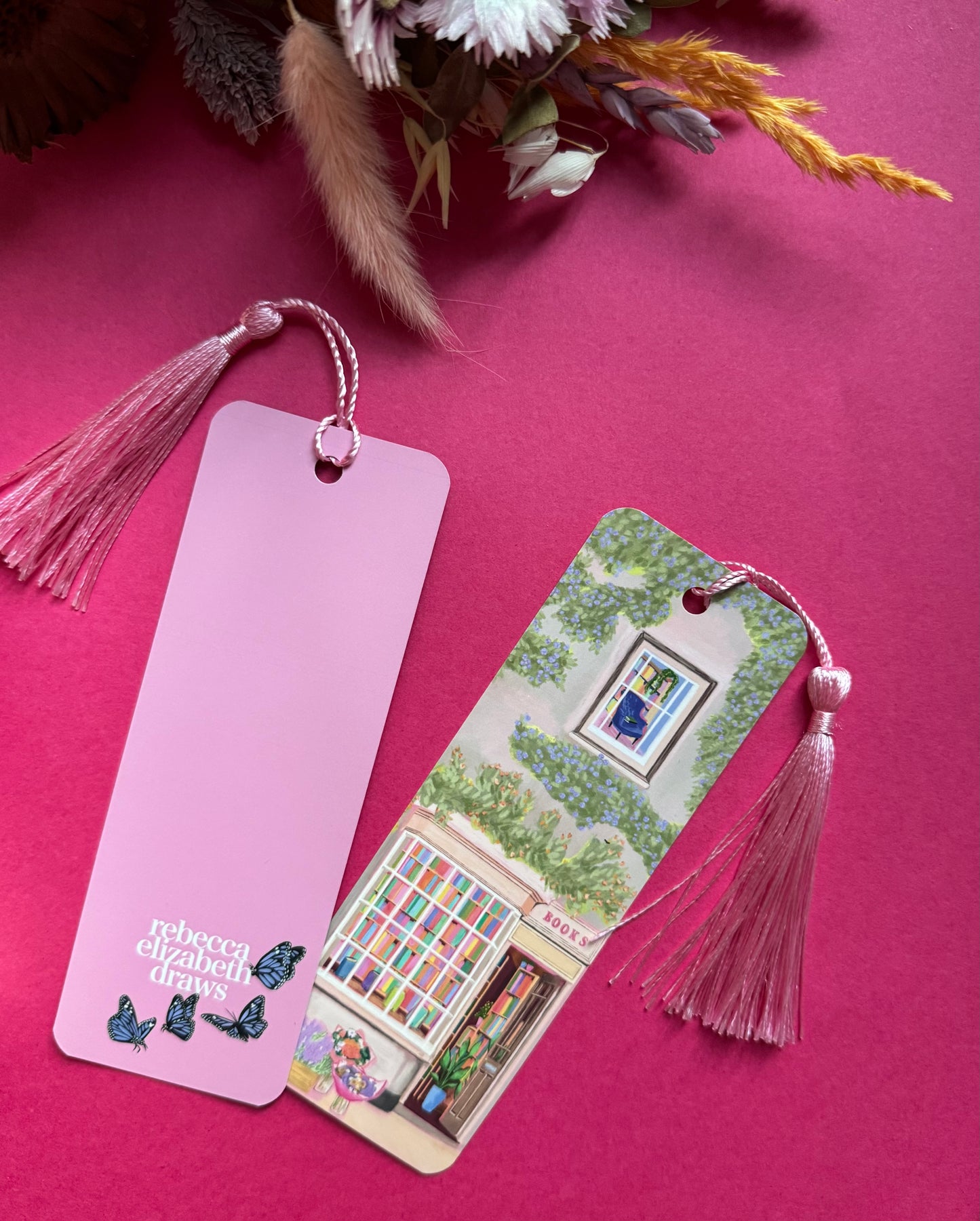 An illustrated bookmark of a cute cosy bookshop, the window shows the bookshelves inside full of bright coloured book spines. There are books placed stood up at the front. Outside the building is a soft colour with lots of greenery in soft pale greens and orange and lilac flowers. In front of the building there are plant pots and flowers. A whimsical cosy art bookmark. There is a small signature in white the corner. Designed by Rebecca Gibbs at Rebecca Elizabeth Draws.