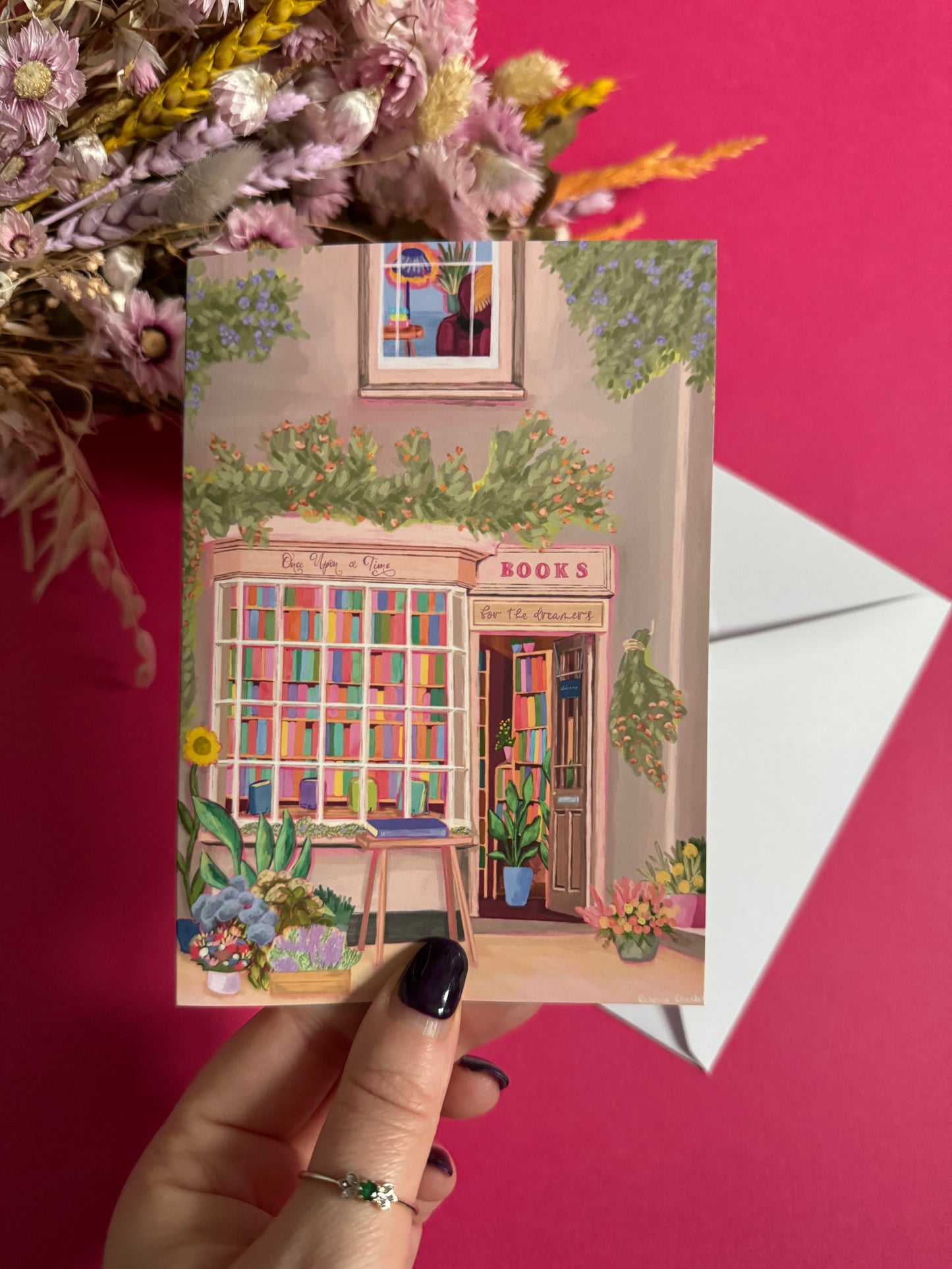 An illustration of a cute whimsical bookshop, the window shows the bookshelves inside full of bright coloured book spines with lots of flowers. The back of the card is pink with the Rebecca Elizabeth Draws logo.