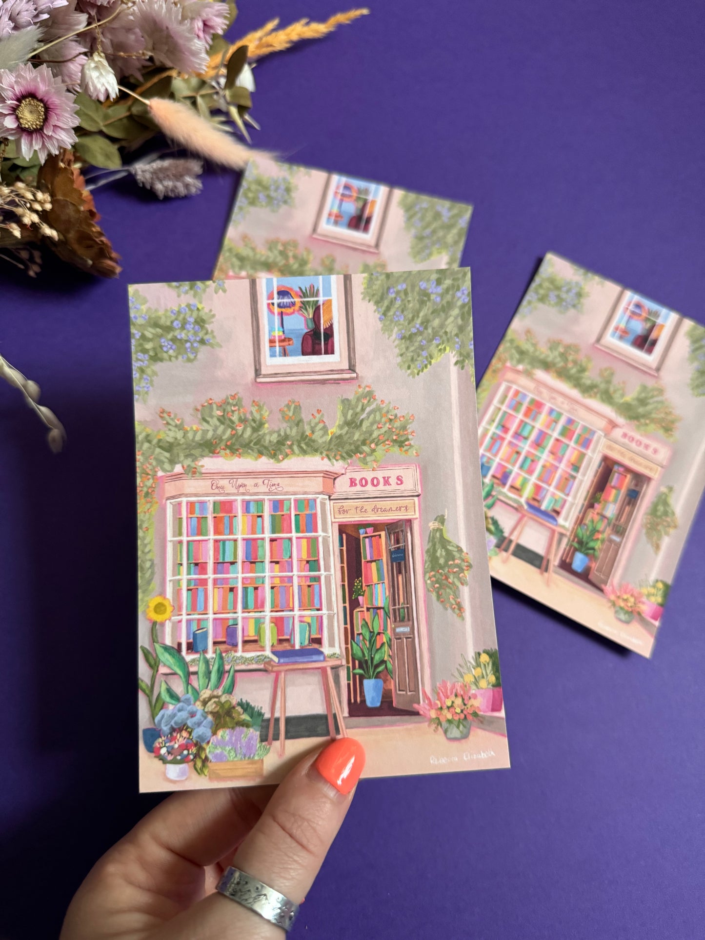 An illustration of a cute bookshop, the window shows the bookshelves inside full of bright coloured book spines. There are books placed stood up at the front. Outside the building is a soft colour with lots of greenery in soft pale greens and orange and lilac flowers. In front of the building there are plant pots and flowers. There is a small signature in white the corner. Designed by Rebecca Gibbs on a postcard at Rebecca Elizabeth Draws.