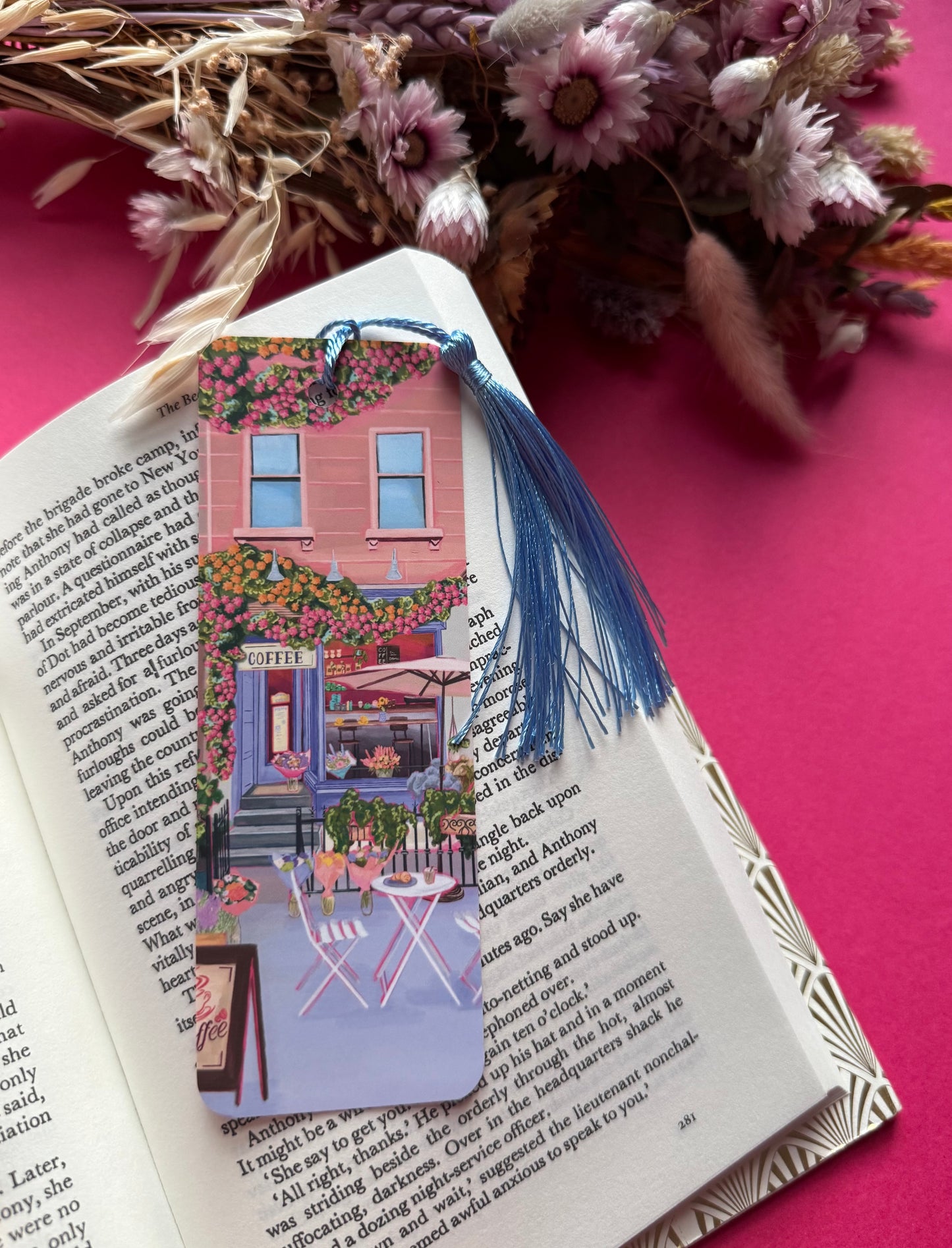 A coffee shop illustrated bookmark with a blue exterior, an outside table and umbrella and building windows surrounding. This illustration is filled with lots of plants and flowers and features cups of coffee and baked goods. Illustrated by Rebecca Elizabeth Draws.
