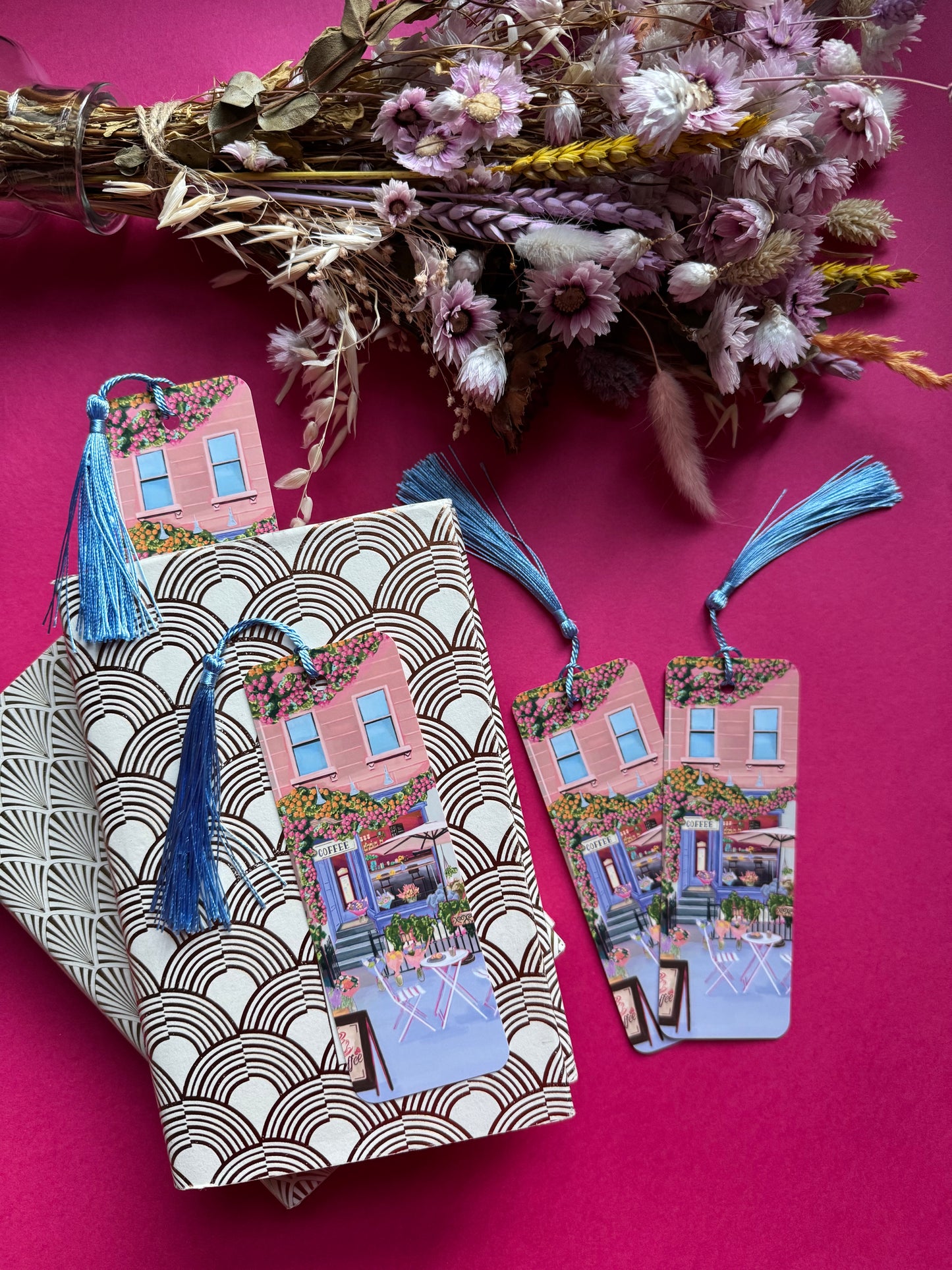 A coffee shop illustrated bookmark with a blue exterior, an outside table and umbrella and building windows surrounding. This illustration is filled with lots of plants and flowers and features cups of coffee and baked goods. Illustrated by Rebecca Elizabeth Draws.