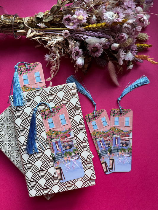 A coffee shop illustrated bookmark with a blue exterior, an outside table and umbrella and building windows surrounding. This illustration is filled with lots of plants and flowers and features cups of coffee and baked goods. Illustrated by Rebecca Elizabeth Draws.