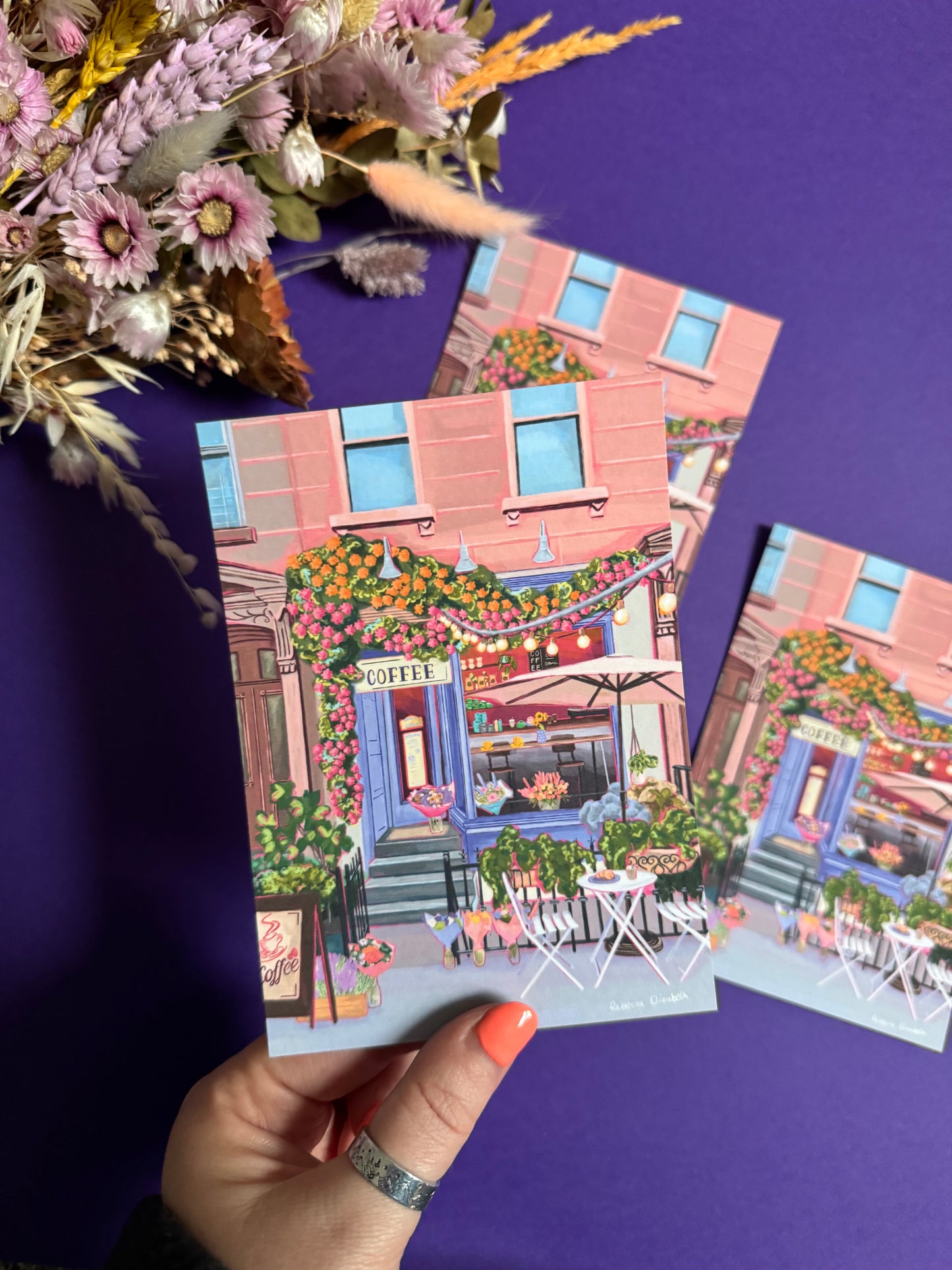 A coffee shop illustration with a blue exterior, an outside table and umbrella and building windows surrounding. This illustration is filled with lots of plants and flowers and features cups of coffee and baked goods. Illustrated by Rebecca Elizabeth Draws.